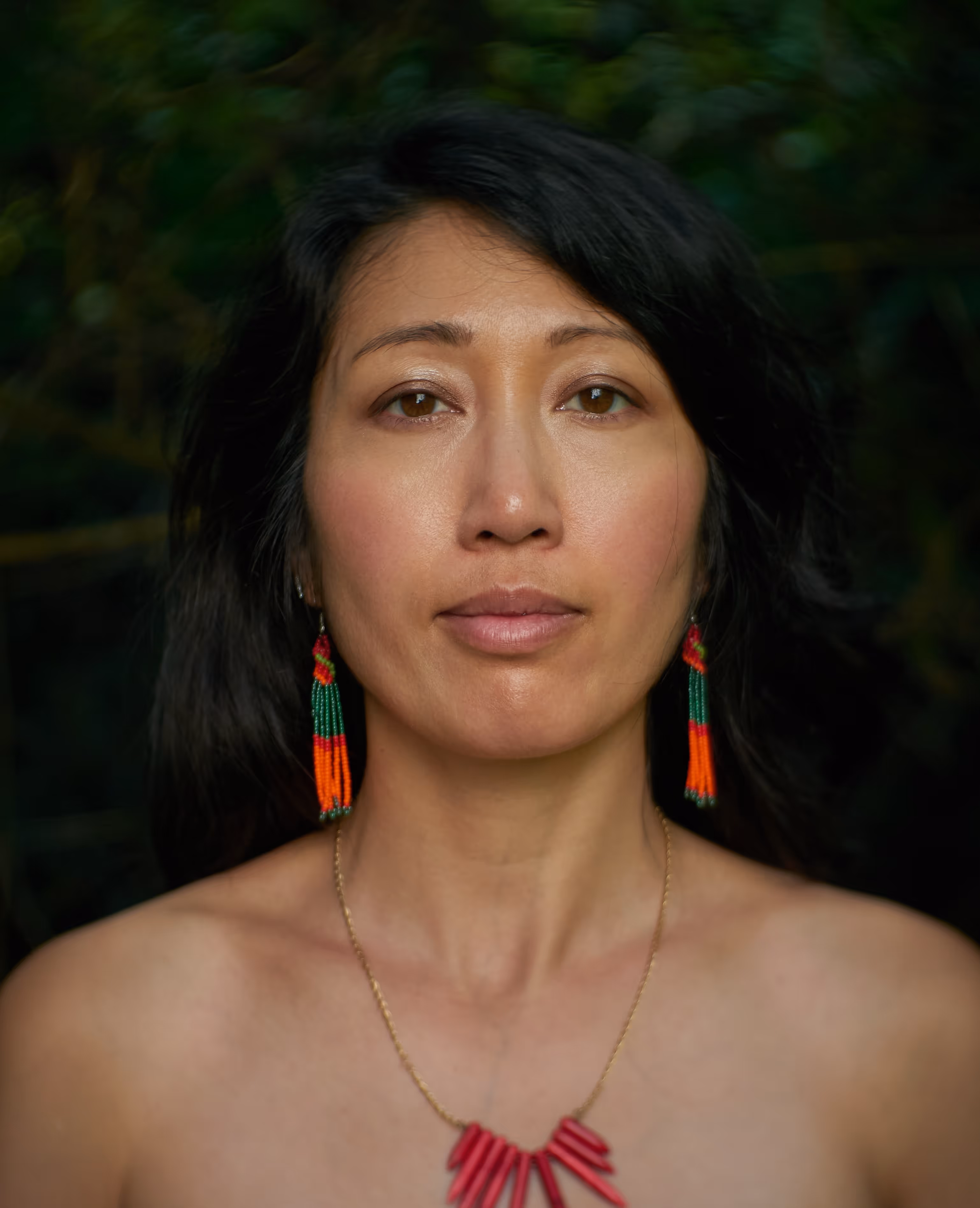 A woman with long dark hair stands against a dark green natural background, wearing vibrant red, orange, and green beaded earrings and a striking red spiked necklace. Her expression is calm and direct, with soft lighting highlighting her face and accessories.