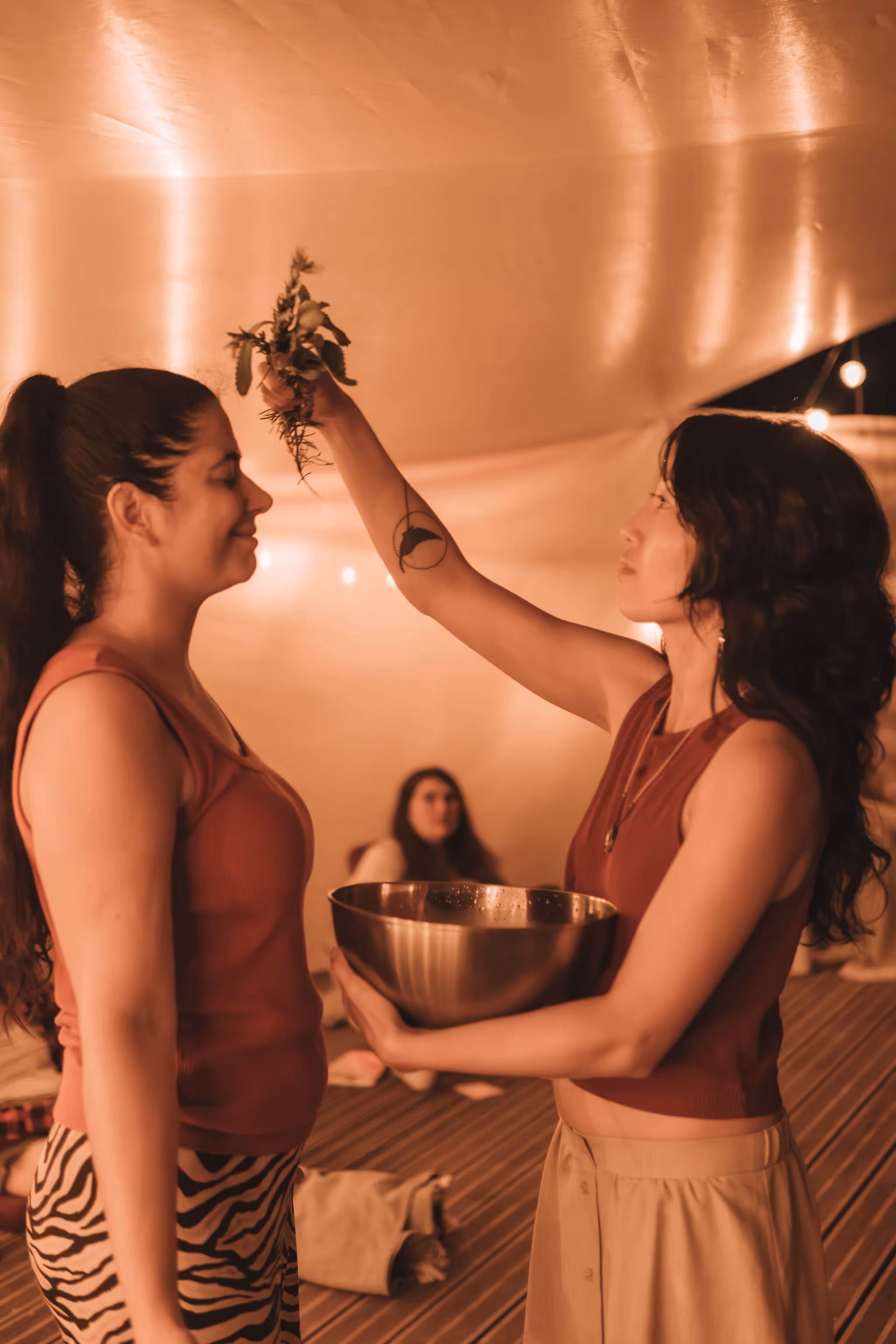 Under warm ambient lighting, a woman holds a metal bowl in one hand while gently raising a bundle of herbs above another woman's head in what appears to be a cleansing or ritual ceremony. Both women are calm and focused, with a third person seated in the background observing the moment.
