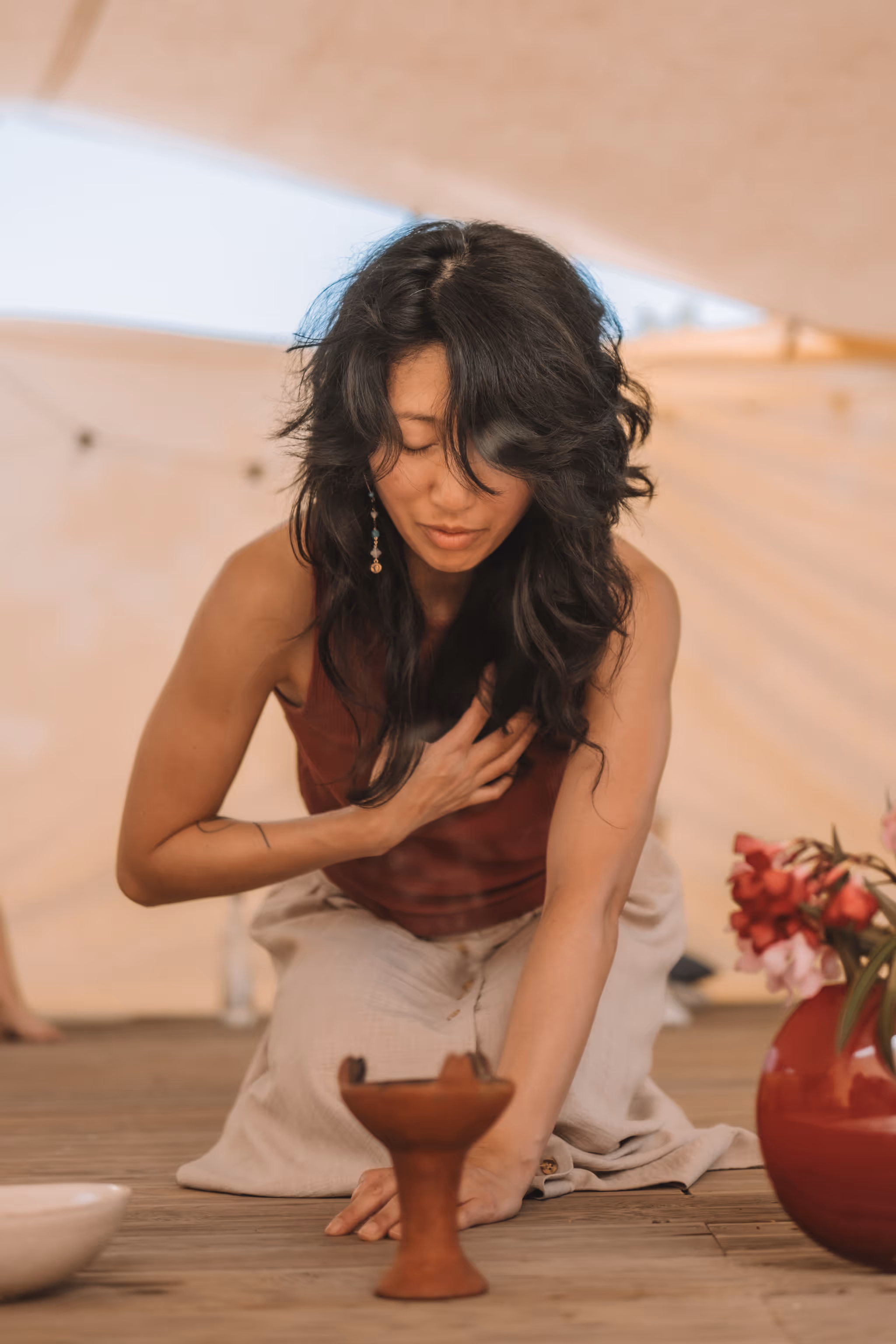 A woman kneels with one hand over her heart and the other resting near a small clay incense burner, eyes gently closed in a moment of intention or prayer. Surrounded by soft light and a vase of pink flowers, the scene evokes ritual, grounding, and self-connection.