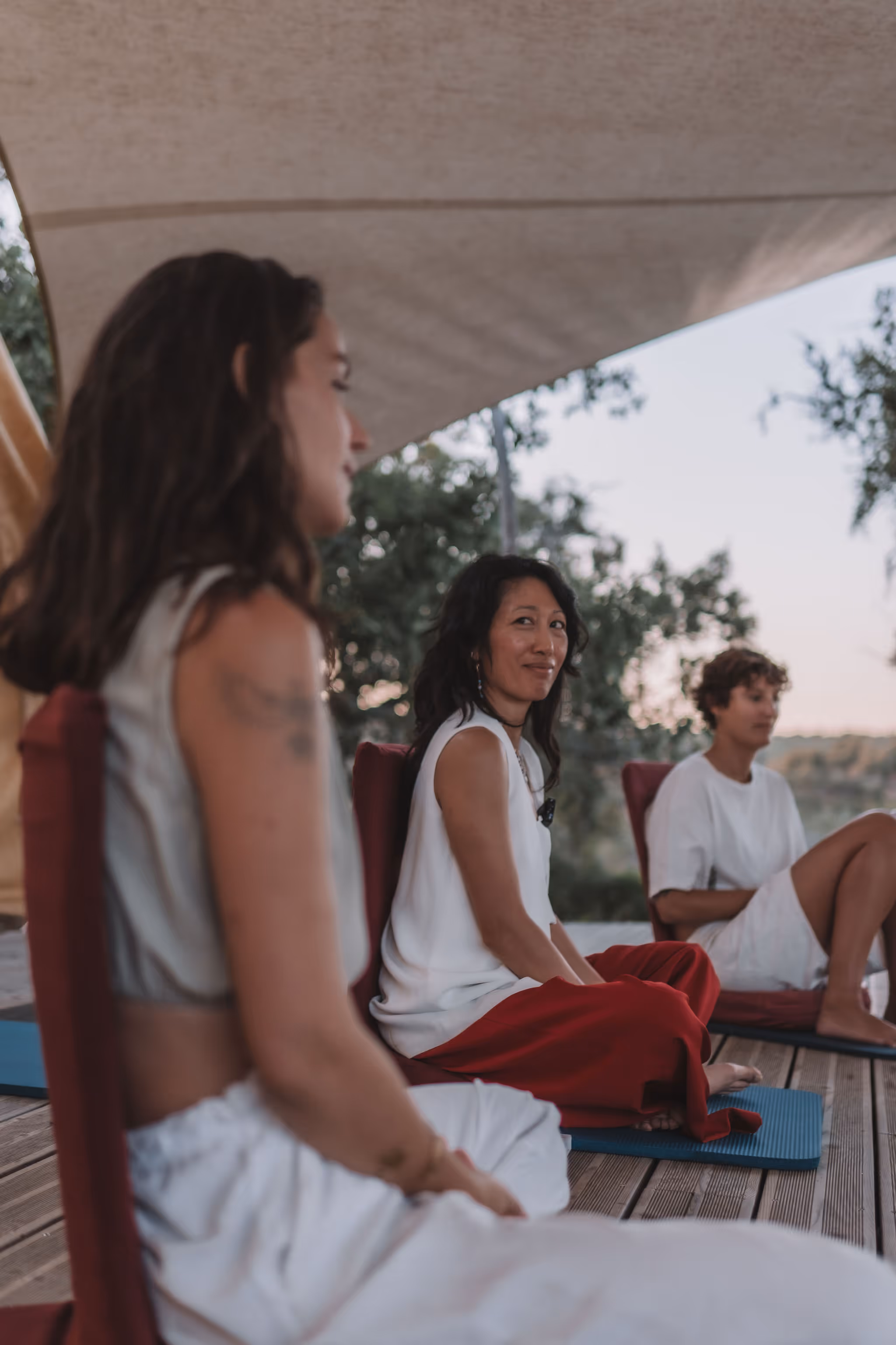 Three women sit cross-legged on mats with backrests under a canopy at dusk, wearing relaxed, light-colored clothing. The central figure smiles gently while turning to look at someone, suggesting a peaceful group discussion or meditation circle.