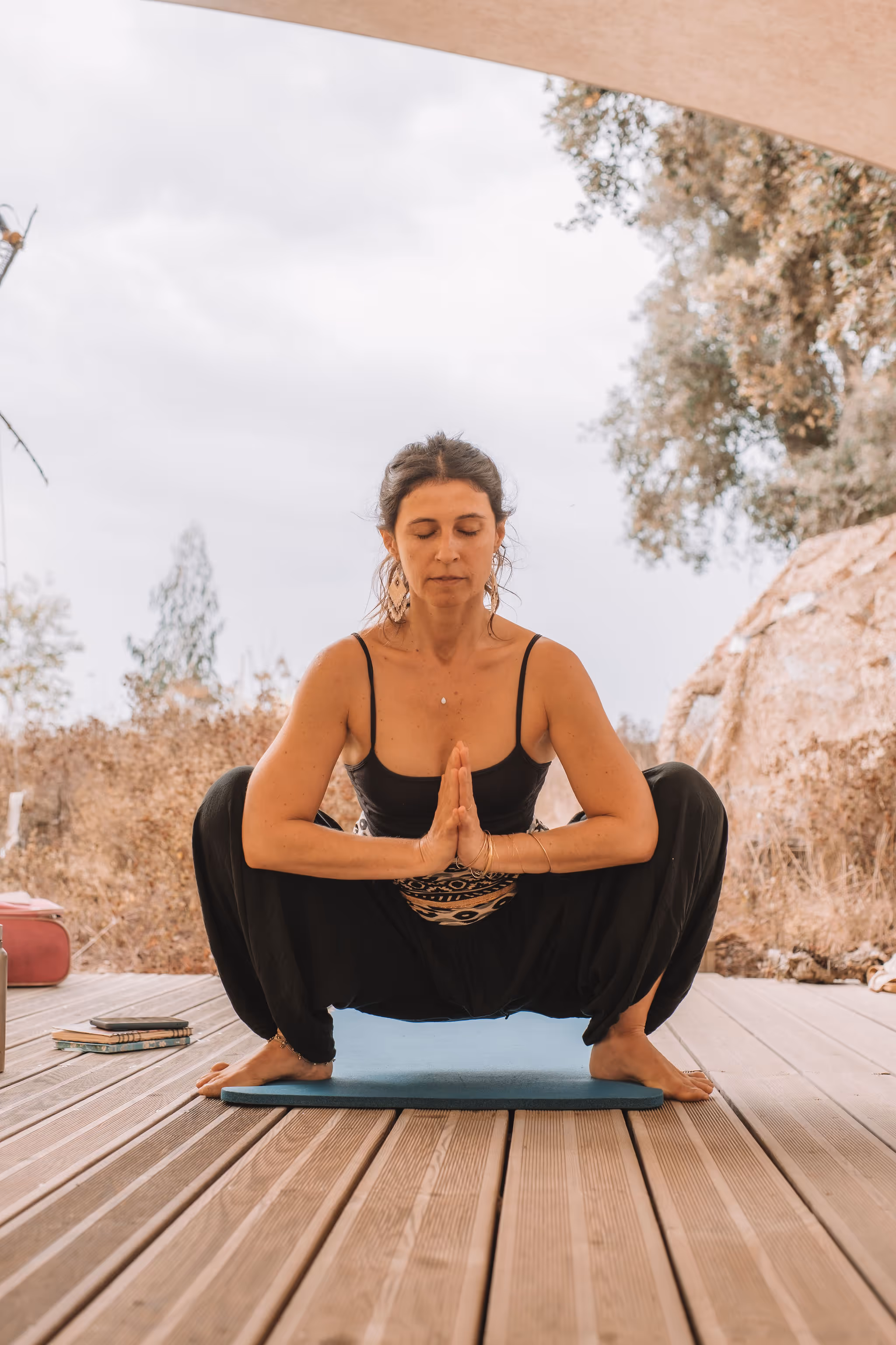 A woman practices a deep squat yoga pose with her hands in prayer position, eyes closed, and a calm expression, seated on a mat under a shaded outdoor area. The peaceful surroundings and focused posture suggest meditation or grounding breathwork in nature.