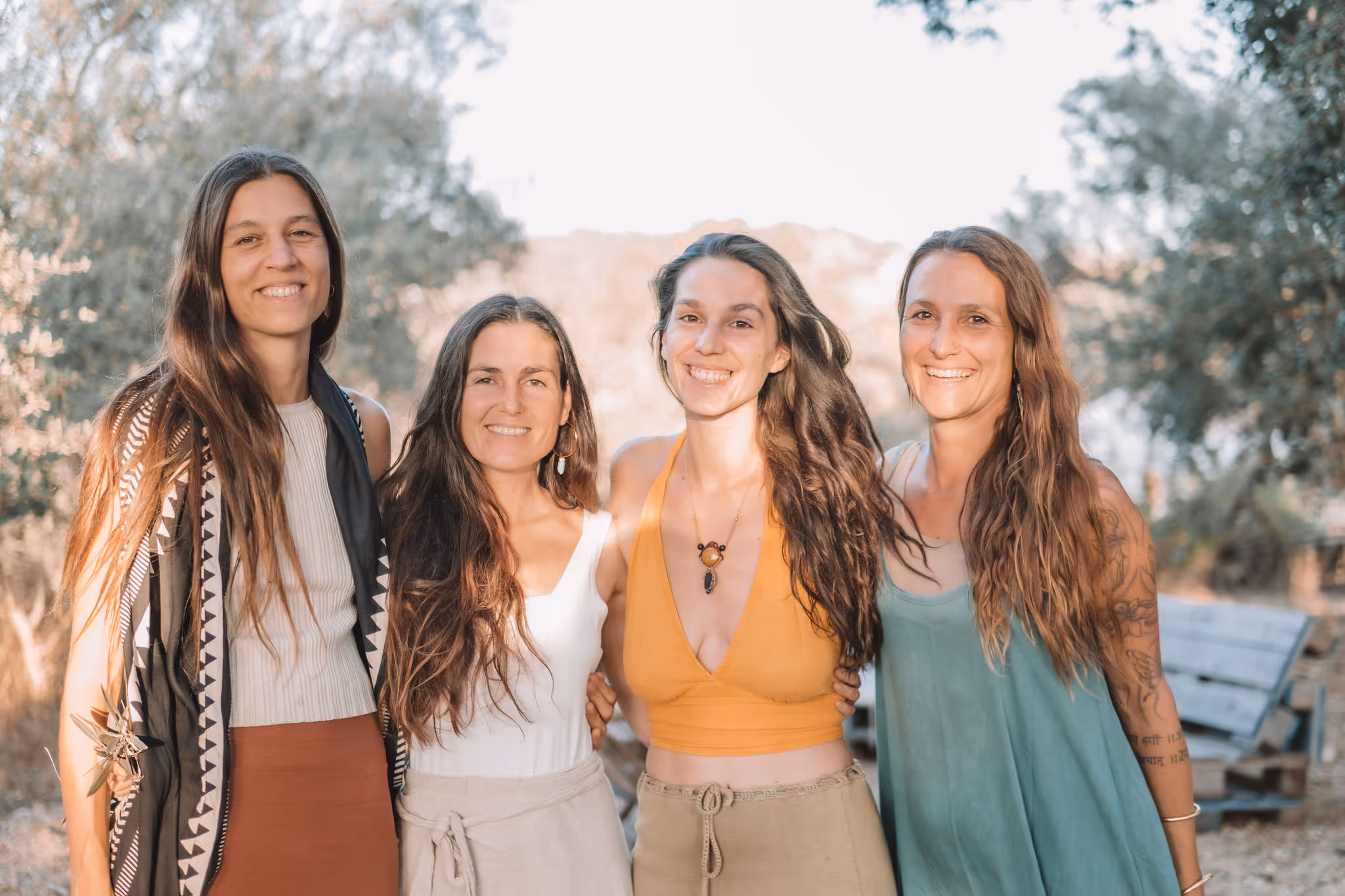 Four facilitators stand closely together outdoors, smiling warmly in soft natural light with trees and hills in the background. They wear relaxed, earthy-toned clothing and have long, flowing hair, suggesting a sense of unity and natural living.
