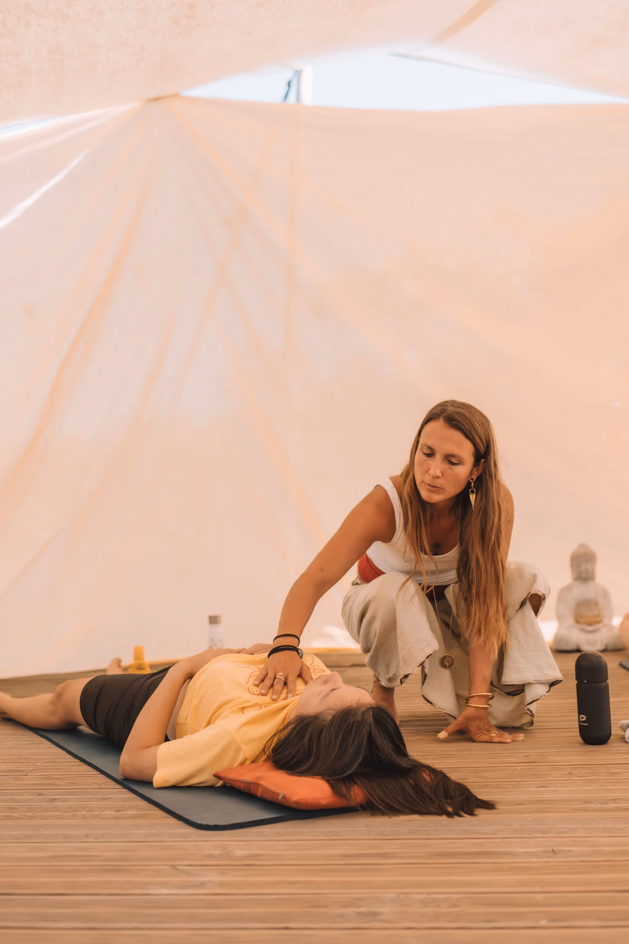 A woman lies on a yoga mat with her eyes closed and hand on her chest, while the retreat facilitator woman gently places her hand on the woman's heart center in a healing gesture. The scene, set under a soft-lit canopy with spiritual decor, evokes a calming, therapeutic energy session or breathwork practice.