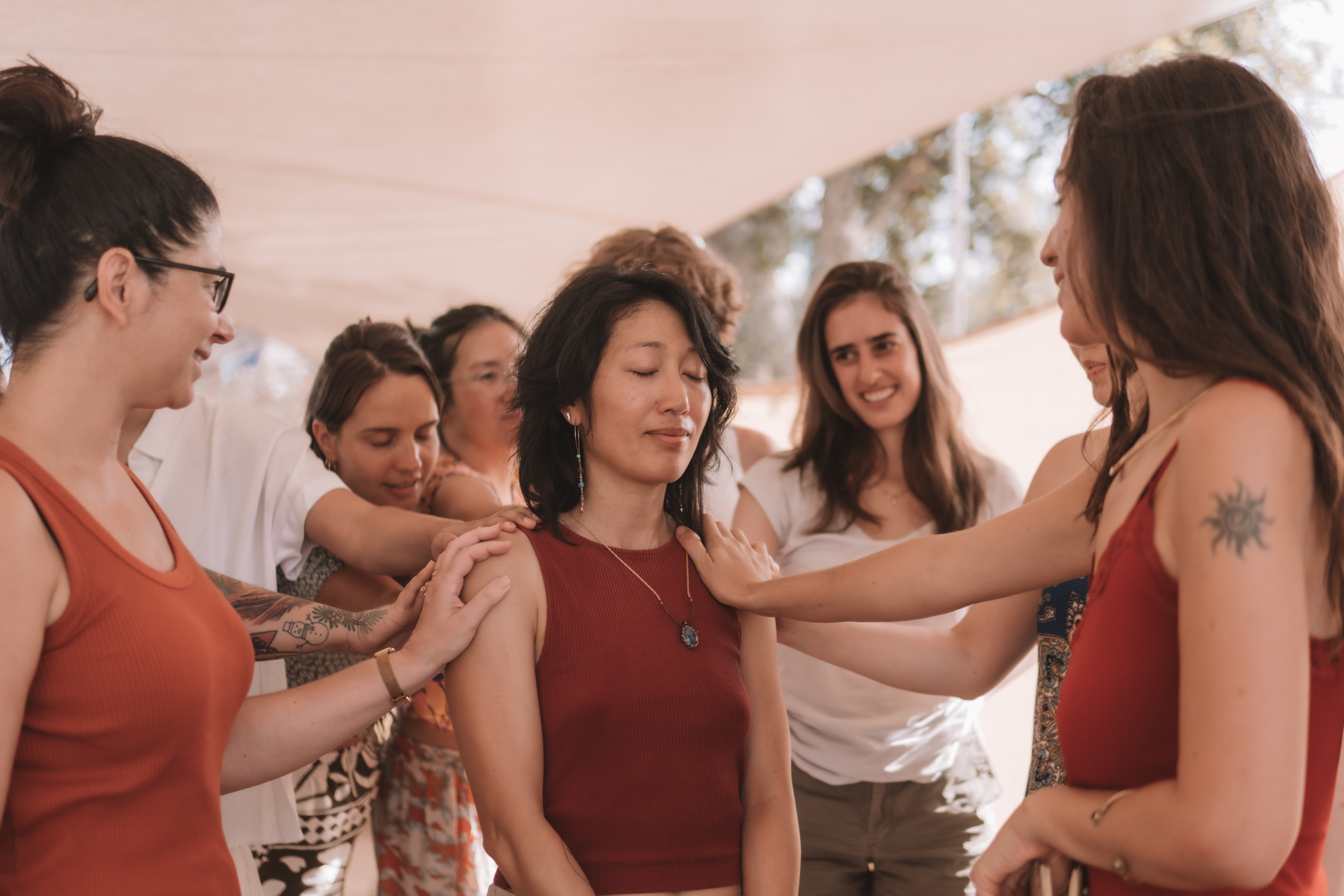 A group of women in casual clothing stands in a circle under a tent, gently placing their hands on one woman's shoulders as she closes her eyes in a calm, meditative expression. The atmosphere is warm and supportive, suggesting a healing or empowerment circle
