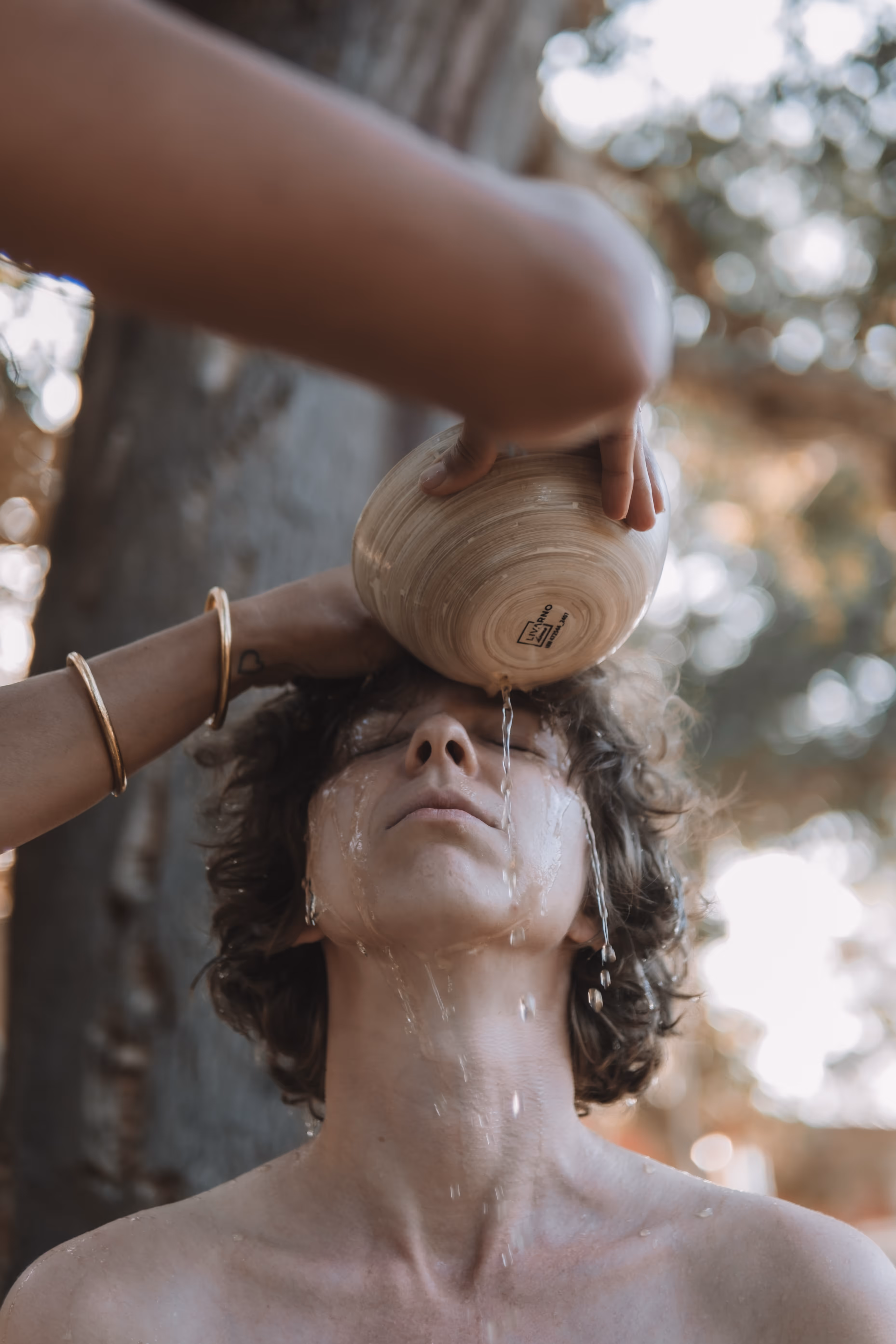 A person with closed eyes tilts their head back as water is gently poured over their forehead from a wooden bowl, streaming down their face and neck. The setting appears to be outdoors, surrounded by trees, suggesting a cleansing ritual or spiritual ceremony.