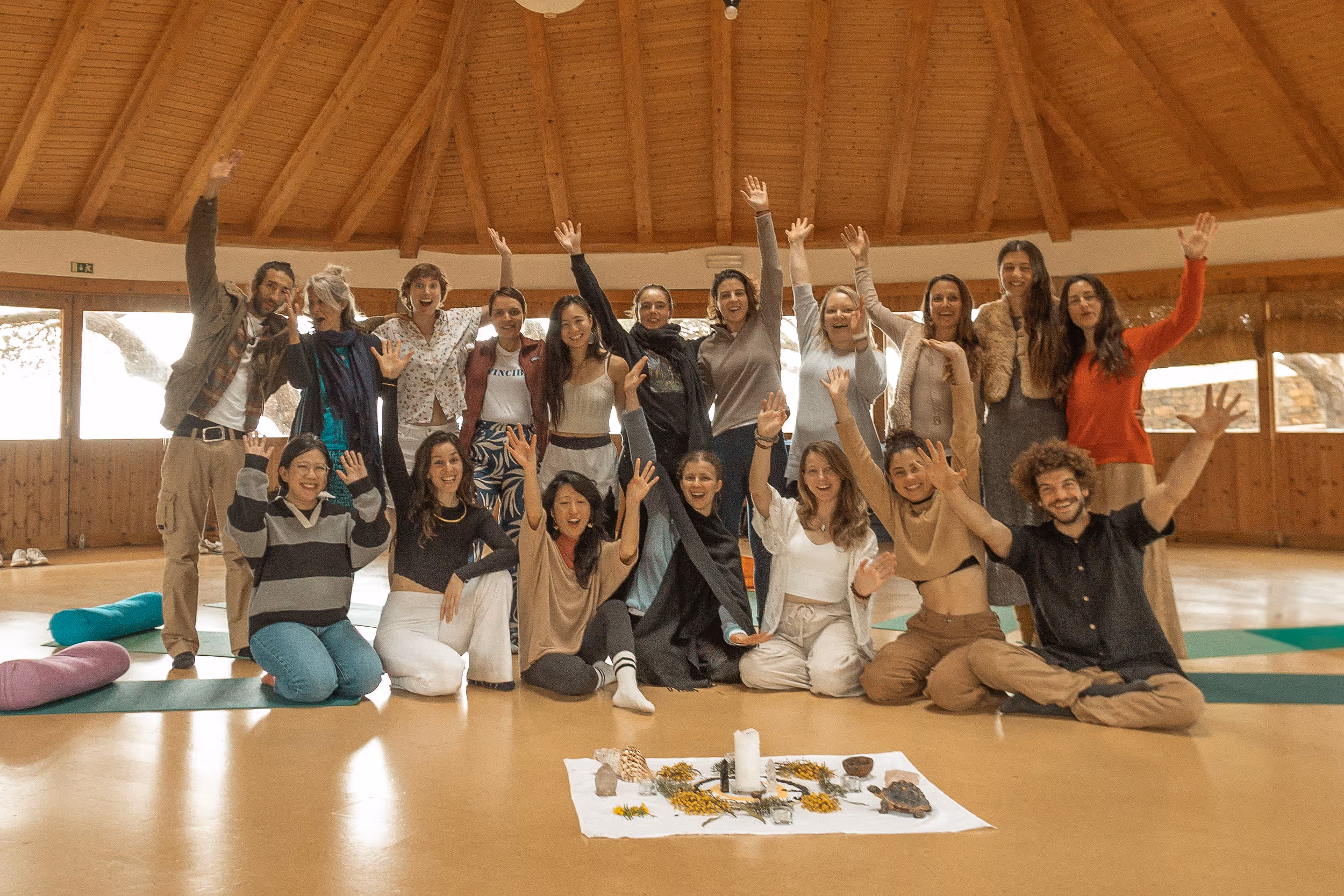 A joyful group of people poses inside a round, wooden-ceilinged room, raising their arms and smiling at the camera. They sit and stand around a central cloth on the floor adorned with candles, flowers, and natural objects, suggesting a wellness or spiritual retreat gathering.
