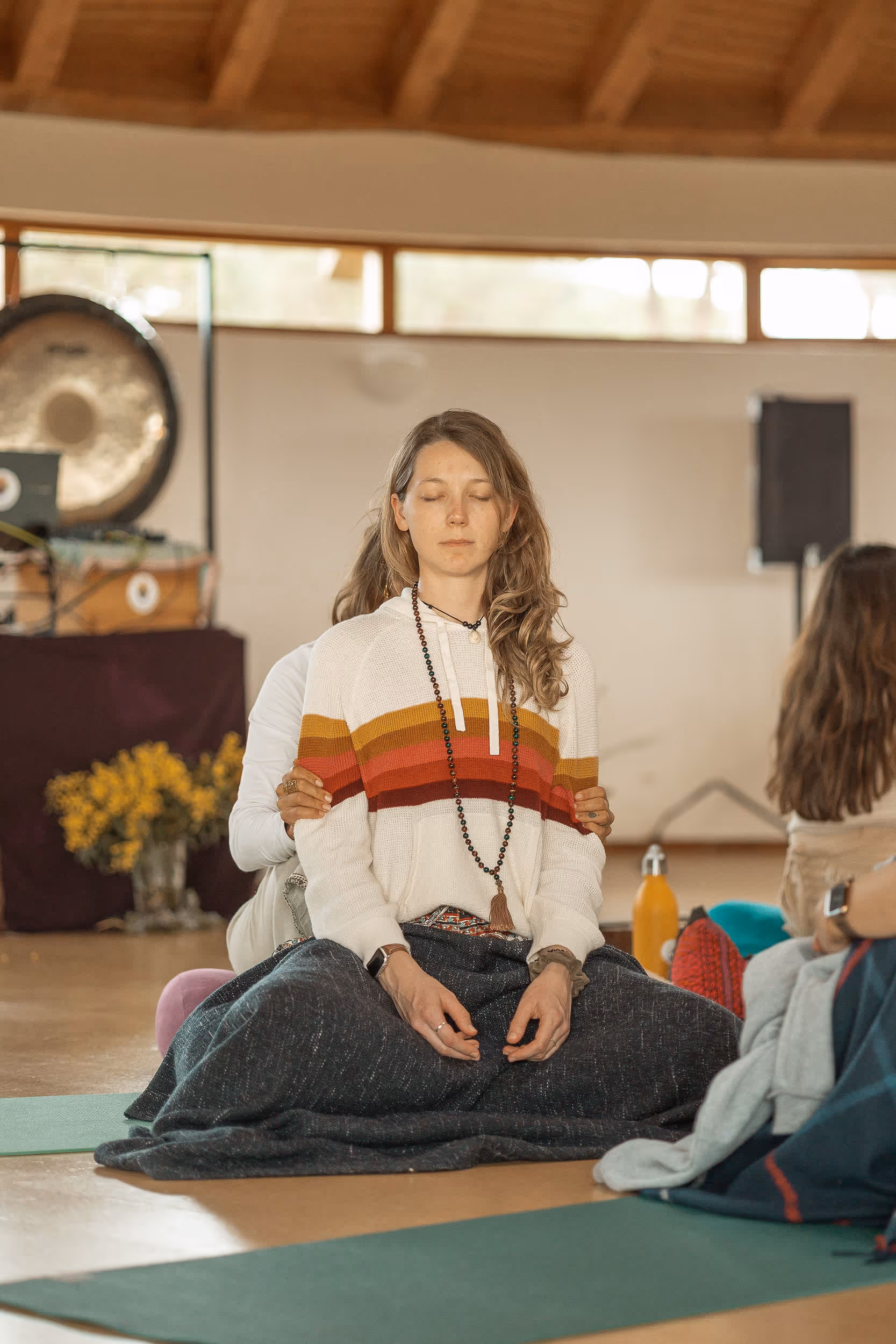A woman sits cross-legged with eyes closed, wrapped in a blanket and wearing a striped sweater and beaded necklace, as another person behind her gently holds her in a grounding or supportive gesture. The setting appears to be a peaceful indoor retreat space, with a gong, flowers, and yoga mats visible in the background.
