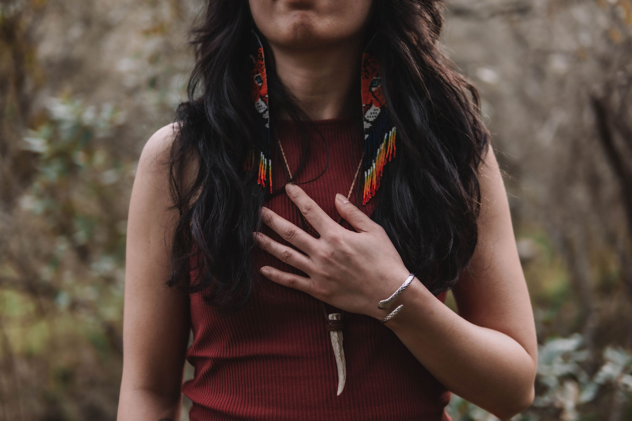 Person wearing a sleeveless burgundy top with long black hair, colorful beaded earrings, a bracelet, and a necklace with a curved pendant, hand resting on chest.