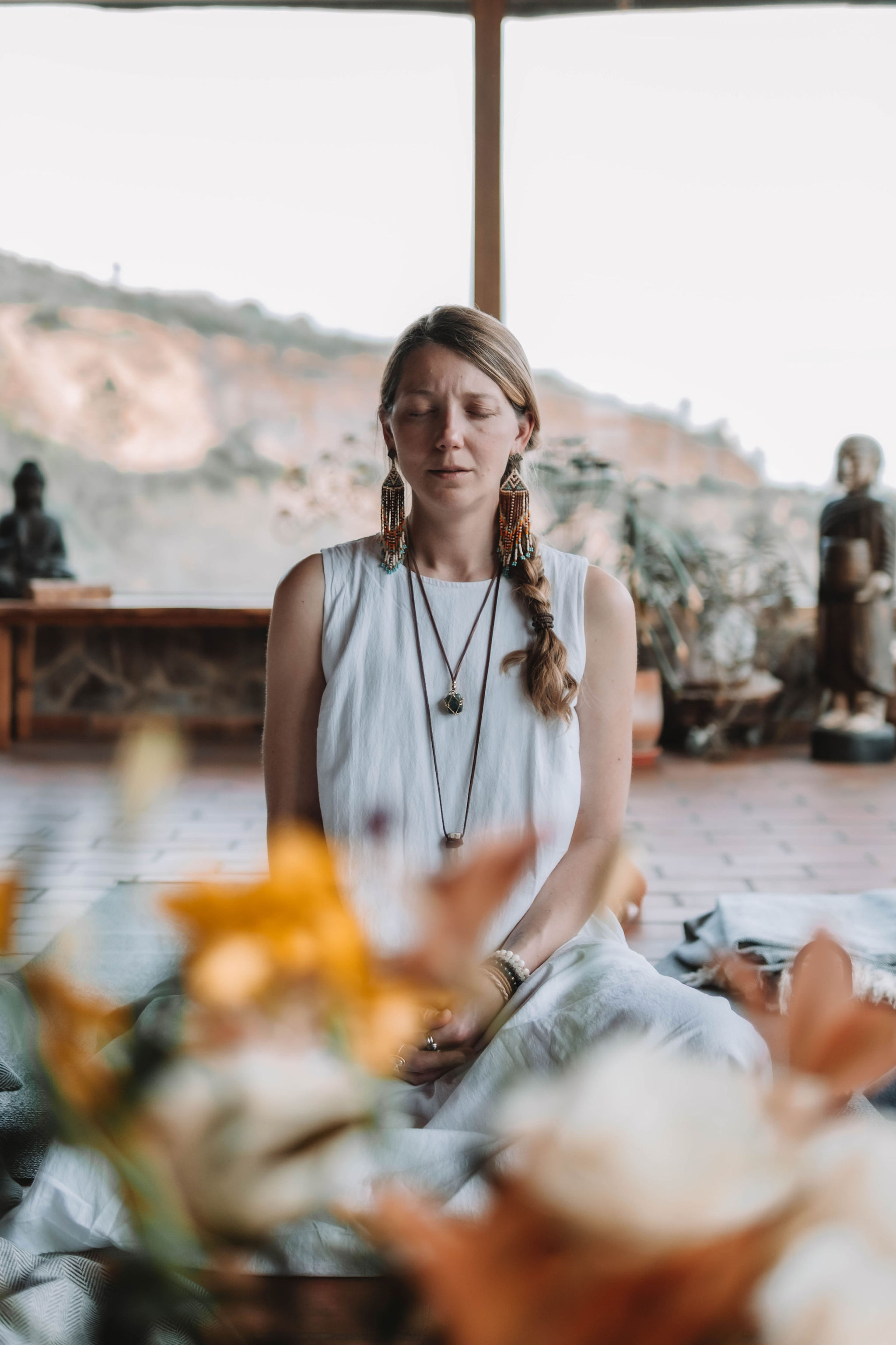Woman seated cross-legged in a white dress meditating with eyes closed in a serene room with blurred flowers in foreground.