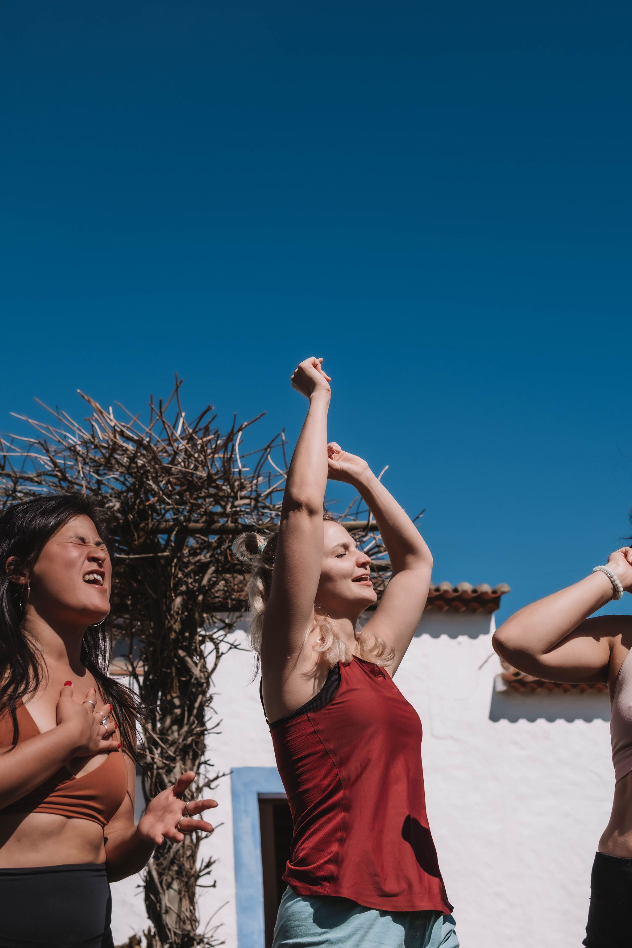 Three women outdoors enjoying a sunny day, appearing joyful and expressive with raised arms and closed eyes under clear blue sky.