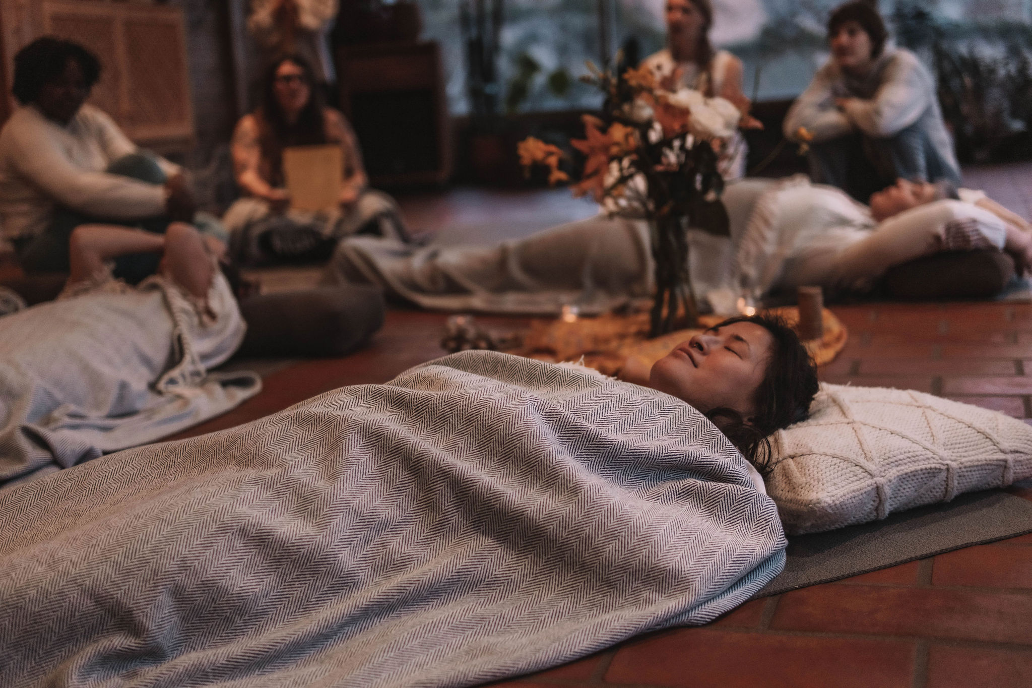 Group of people lying on the floor covered with blankets, relaxing in a cozy indoor setting with flowers and candles in the background.