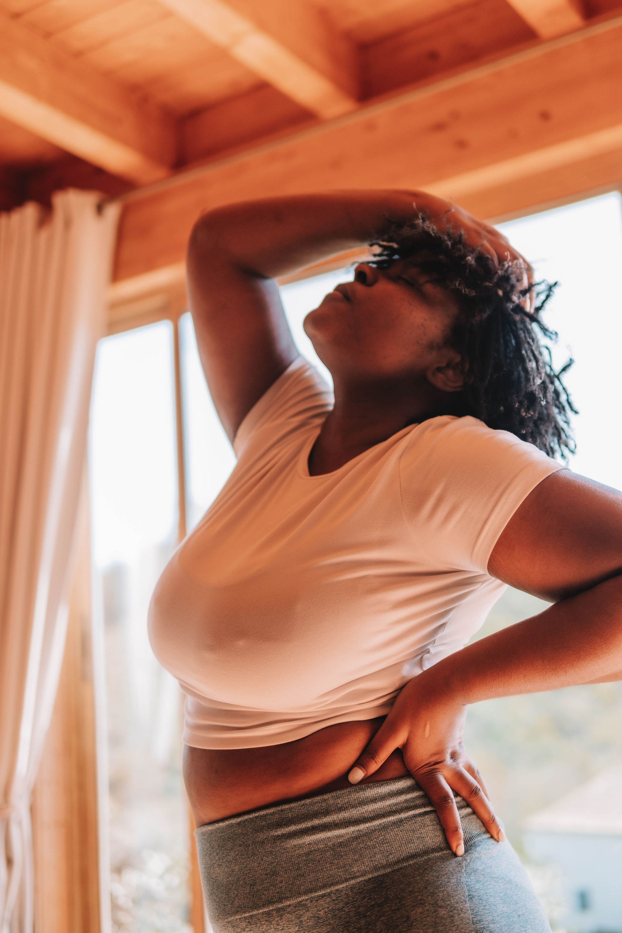 Woman in a white t-shirt and gray pants stretching indoors near a window with a wooden ceiling.