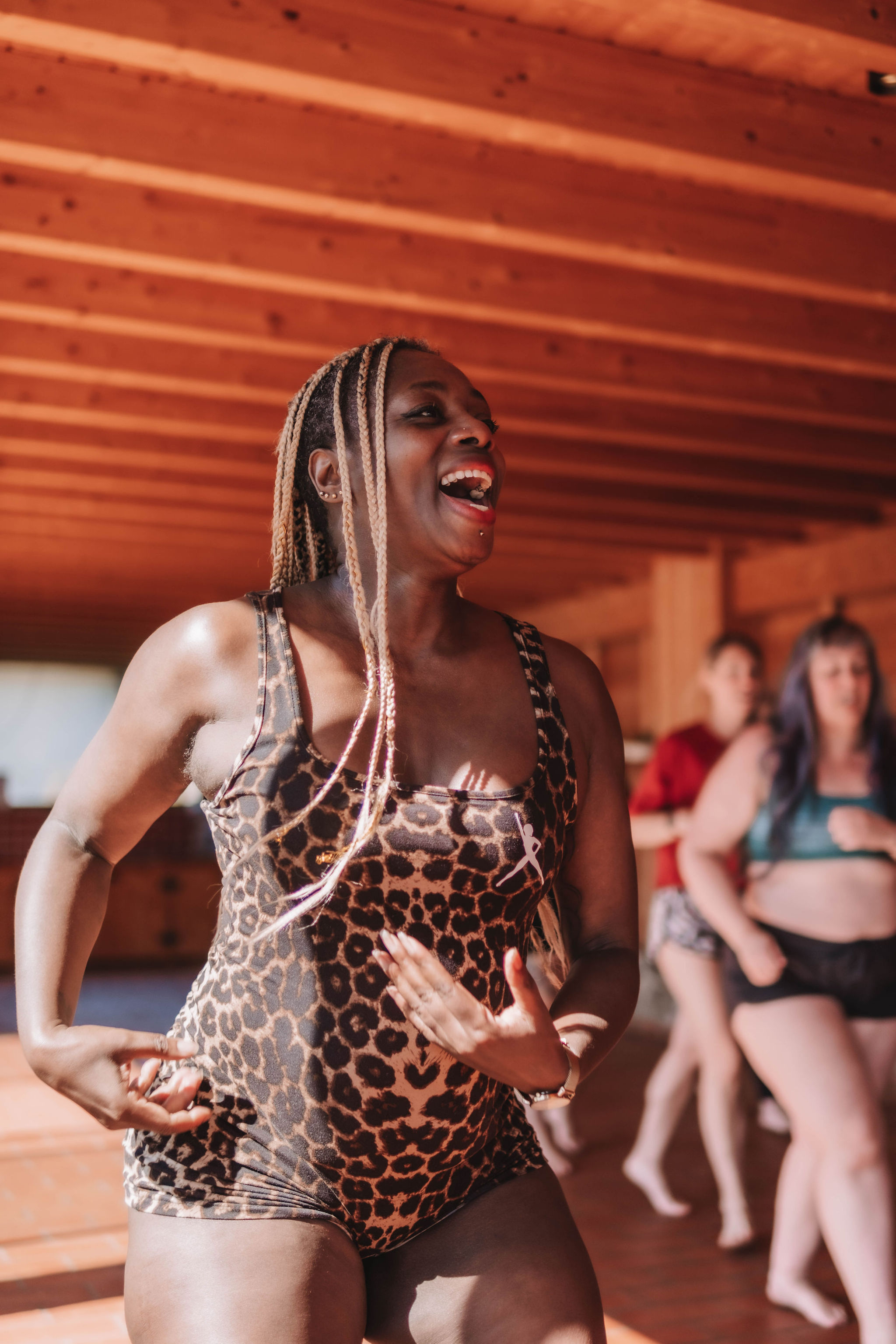 Smiling woman with braided hair in leopard print workout clothes dancing indoors with others in the background.