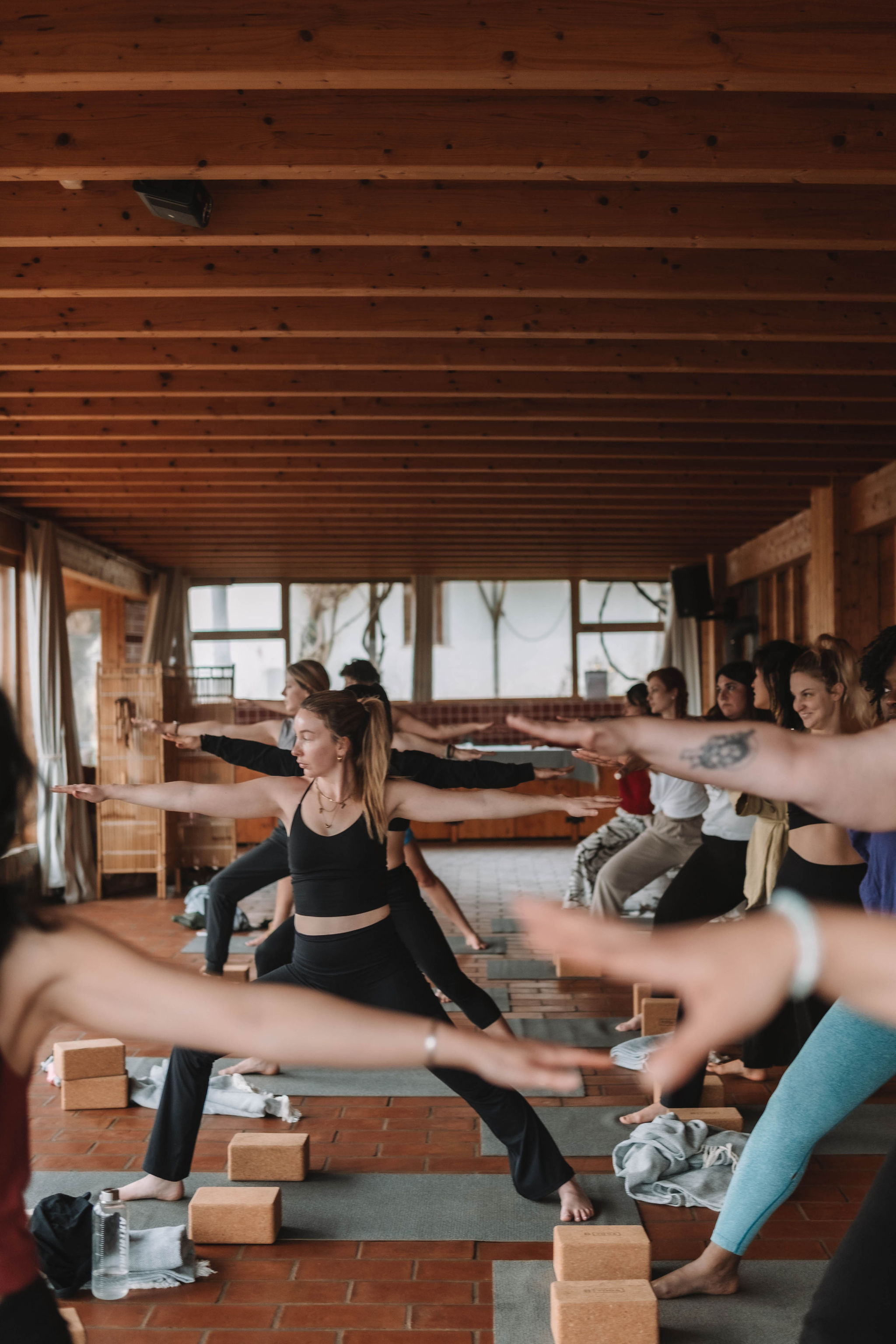 Group of people practicing yoga in warrior pose on mats with yoga blocks in a wooden room.
