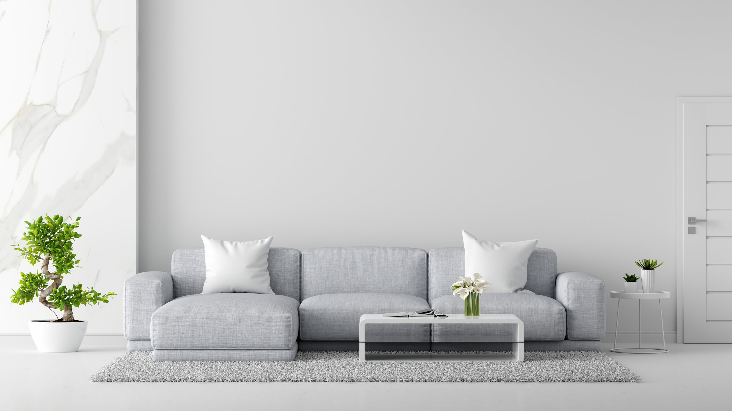 Minimalist living room with a light gray sofa, white pillows, a white coffee table with flowers and glasses, a gray rug, and green plants on both sides.