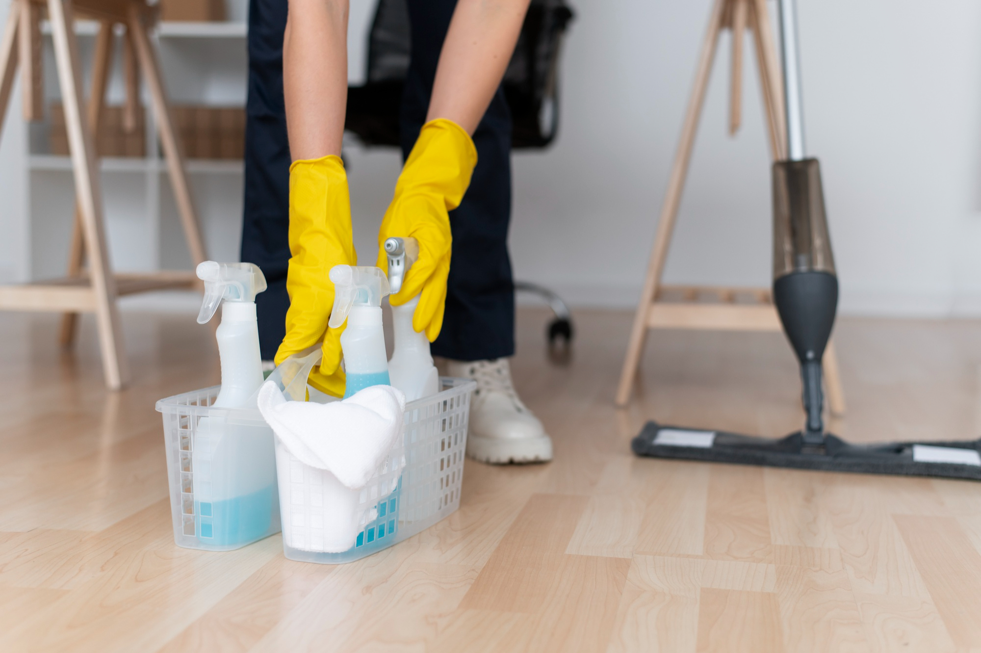 Person wearing yellow gloves holding a basket with cleaning spray bottles and cloth on a wooden floor near a mop.
