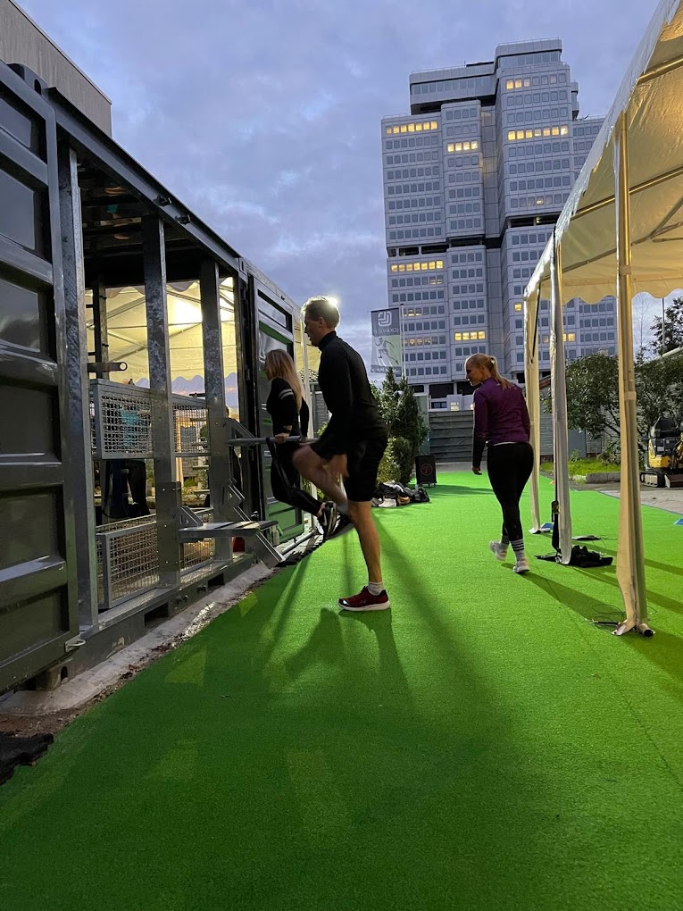 Drei Personen machen Dehnübungen auf grünem Kunstrasen vor einem Containergebäude bei Dämmerung mit Hochhaus im Hintergrund.