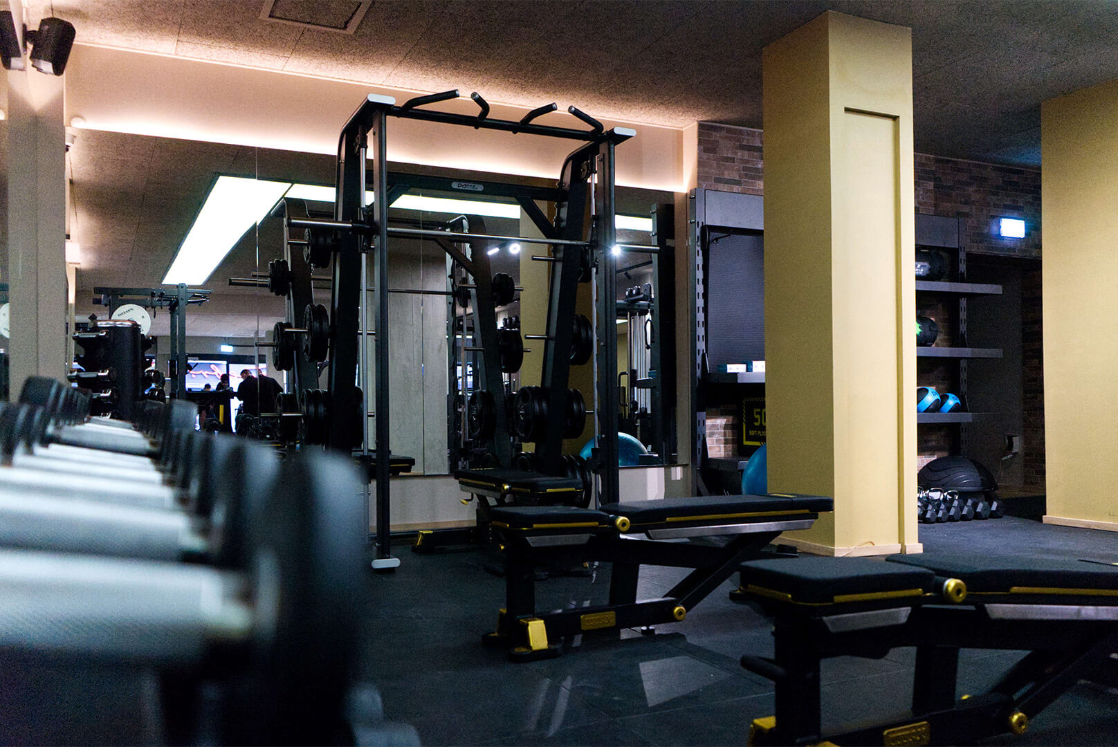 Innenaufnahme eines Fitnessstudios mit Hanteln, Kraftgeräten und Trainingsbänken unter Deckenbeleuchtung.