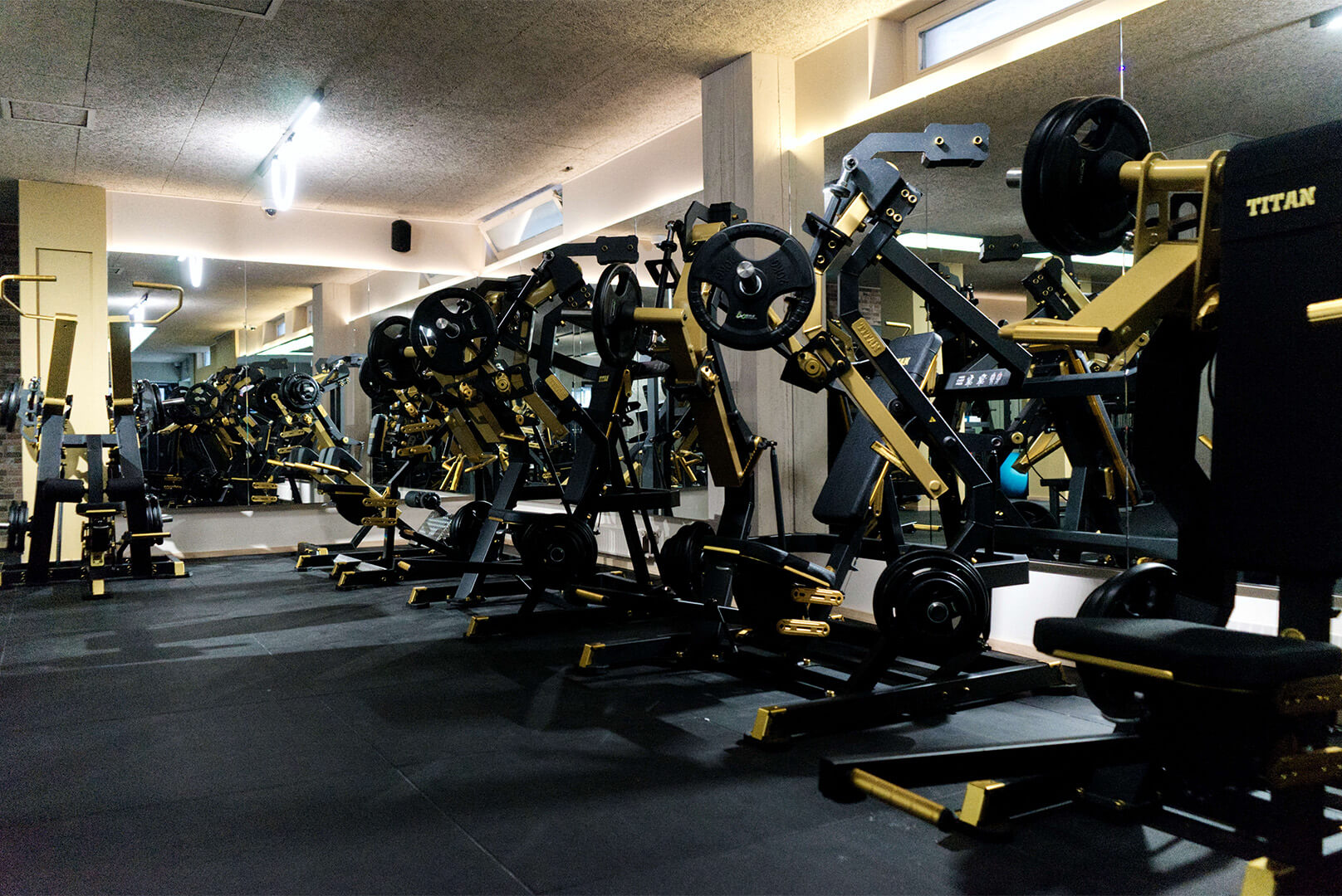Innenansicht eines Fitnessstudios mit schwarzen und goldfarbenen Krafttrainingsgeräten vor großen Spiegeln.