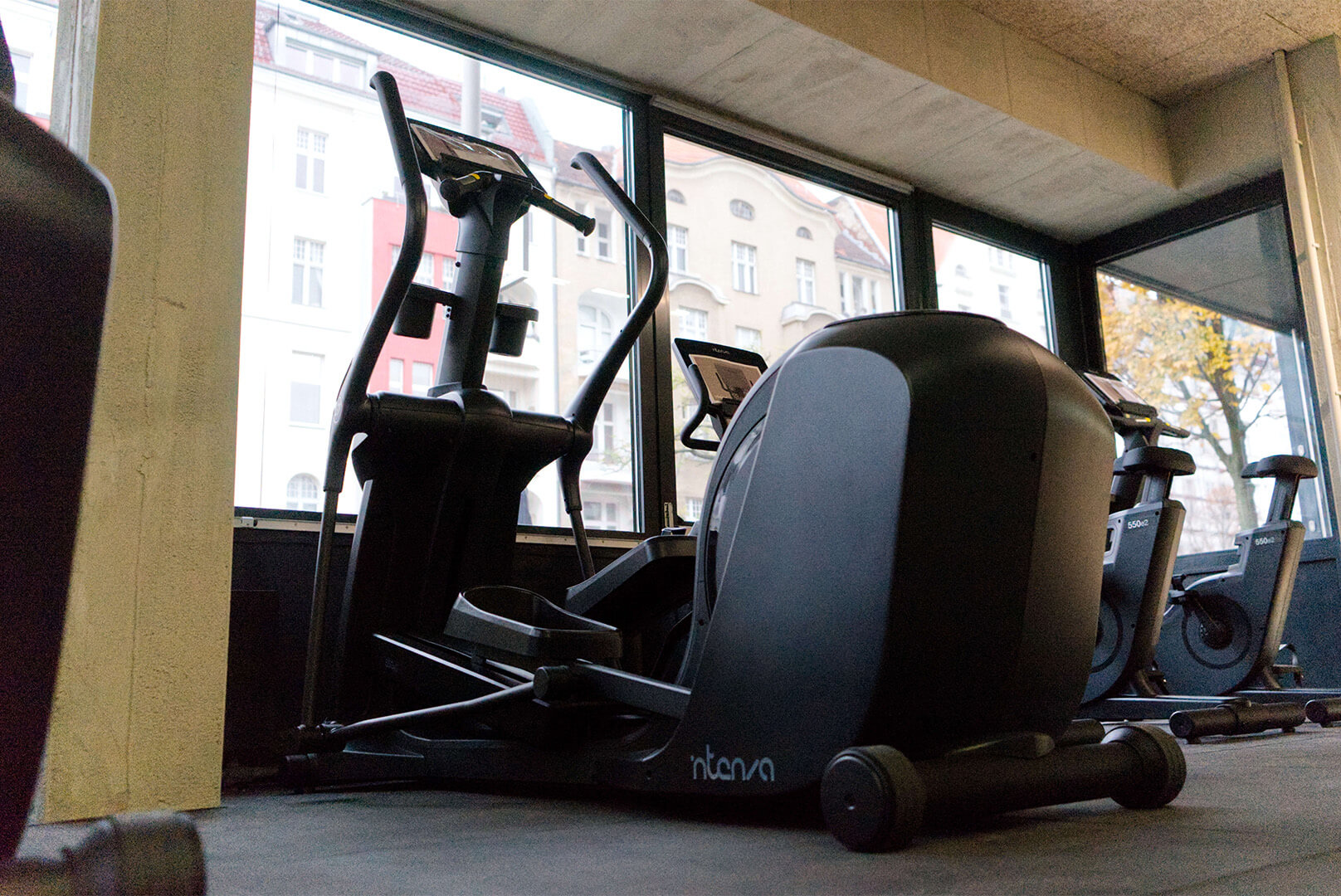 Ellipsentrainer im Fitnessraum vor großen Fenstern mit Blick auf Gebäude und Bäume.