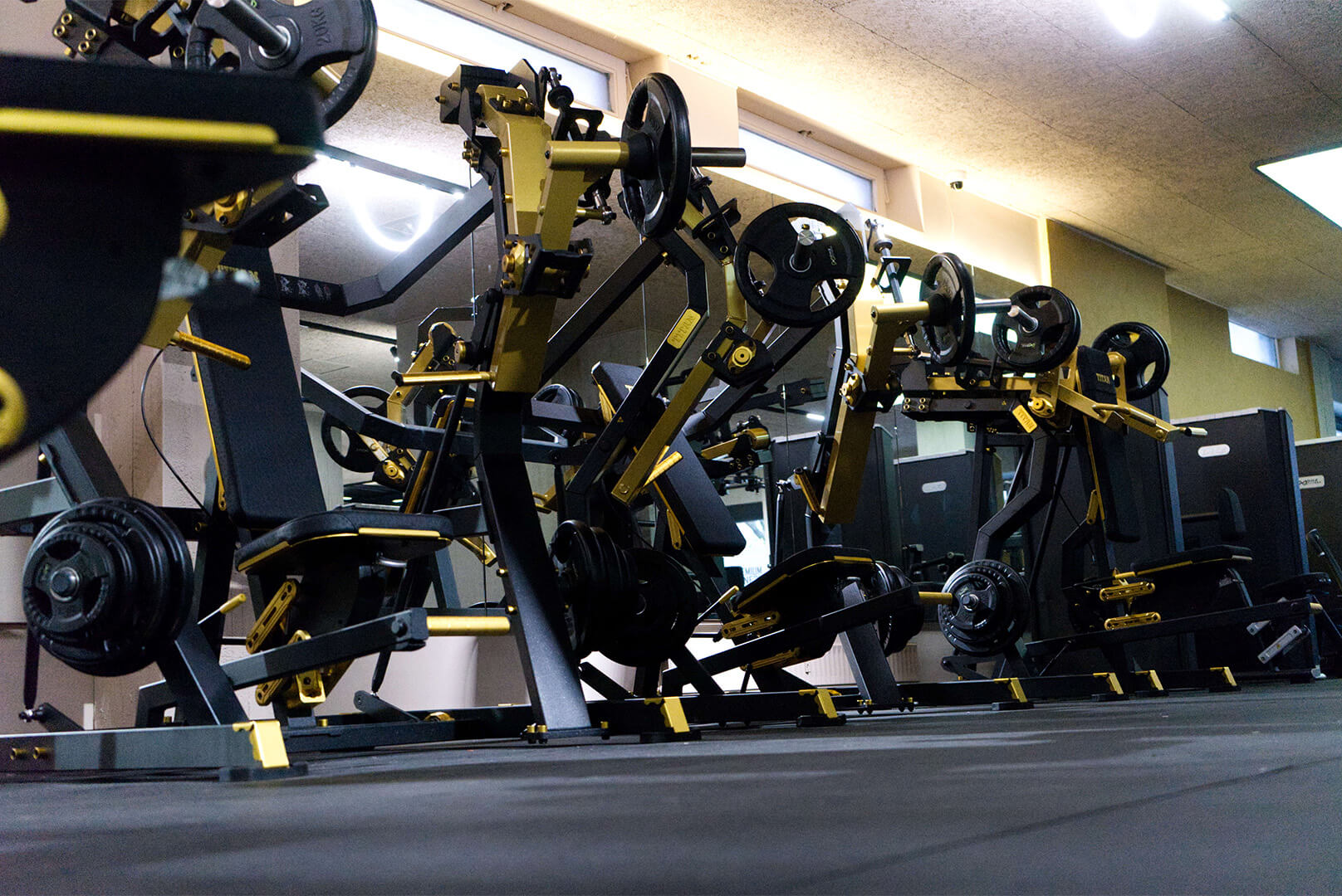 Mehrere Kraftsportgeräte mit schwarzen und goldenen Gewichten in einem Fitnessstudio vor großen Spiegeln.