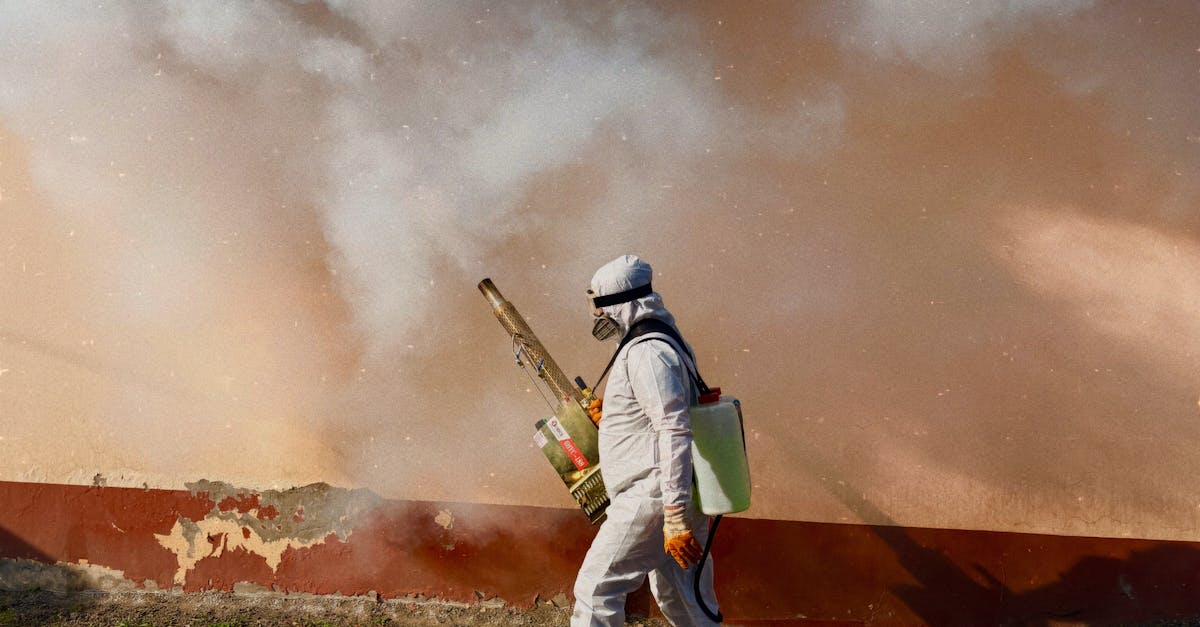 A worker in a full protective suit fumigates an area outdoors, spraying pesticide.