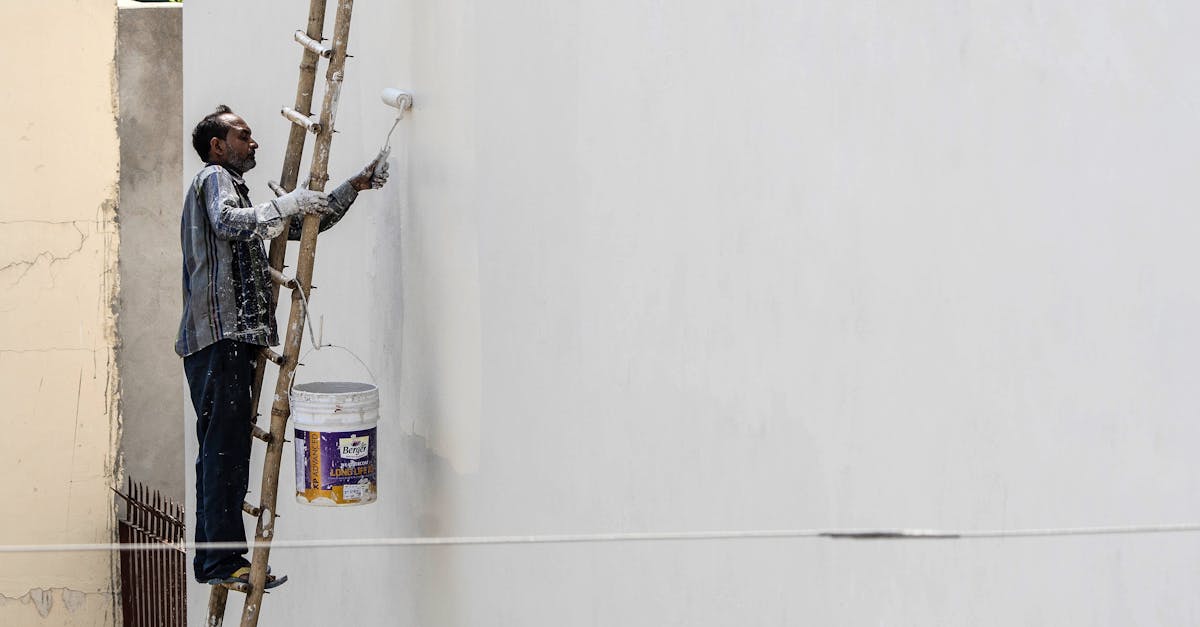 A man on a ladder painting a building's exterior wall with a roller brush outdoors.