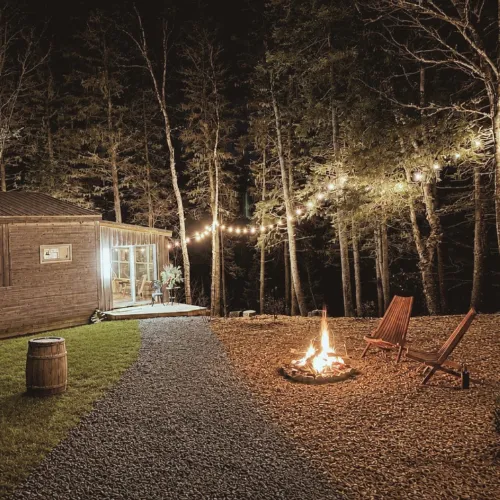 Fire pit glowing at night with Adirondack chairs and string lights in the trees