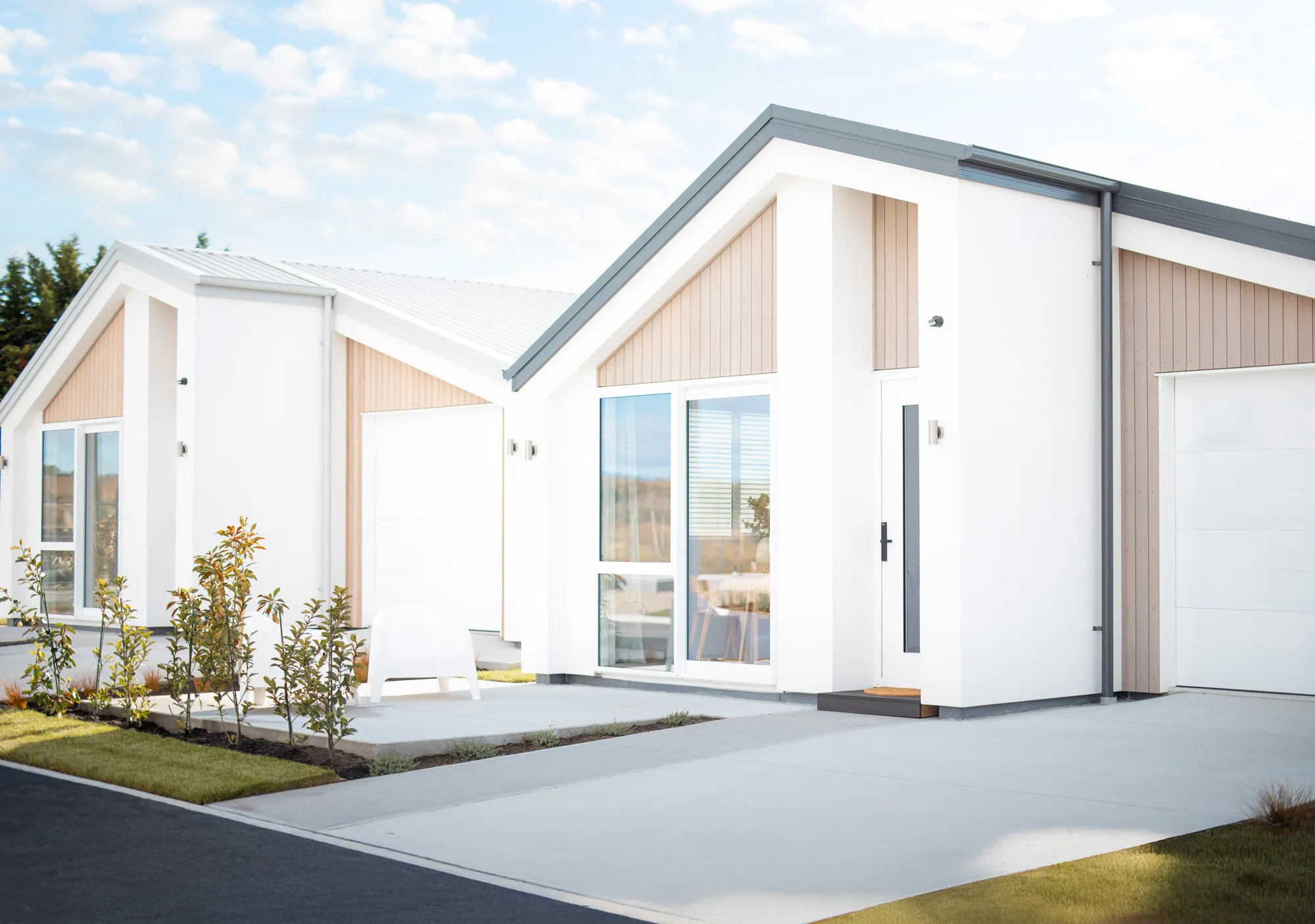 Modern white single-story homes with beige panel accents, large windows, clean concrete driveways, small shrubs, and a white chair on a patio, under a partly cloudy sky.