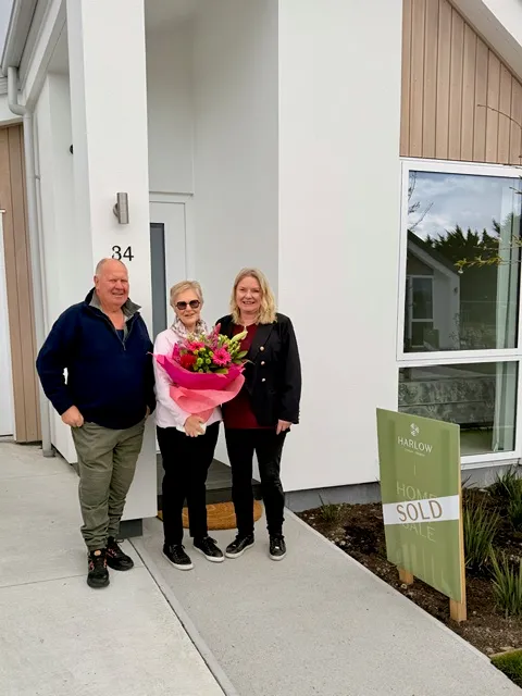 Fiona welcoming new residents outside a there new modern white house with number 34, one person holding a bouquet of flowers, next to a sold sign.