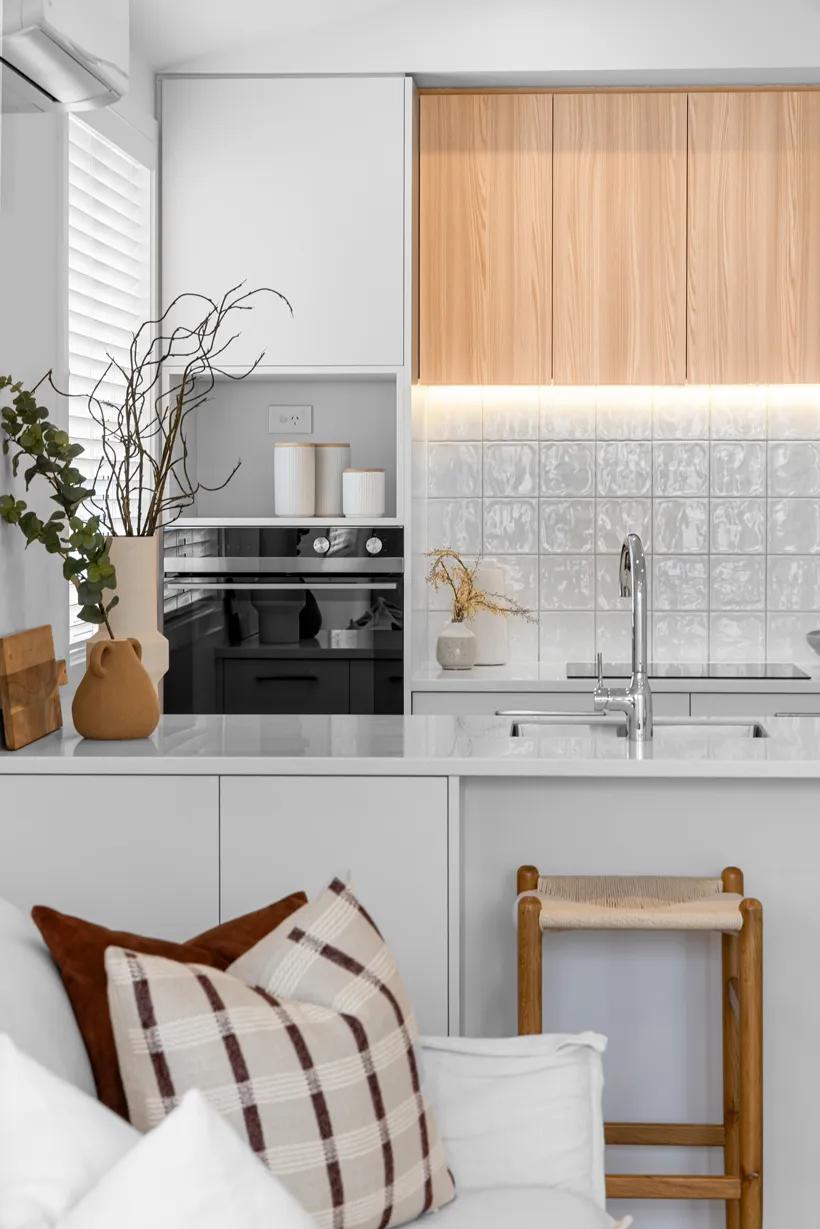 Modern kitchen with white countertop and light wood cabinets, featuring a built-in black oven, chrome faucet, decorative vases, and a beige woven stool with a white sofa and plaid pillows in the foreground.