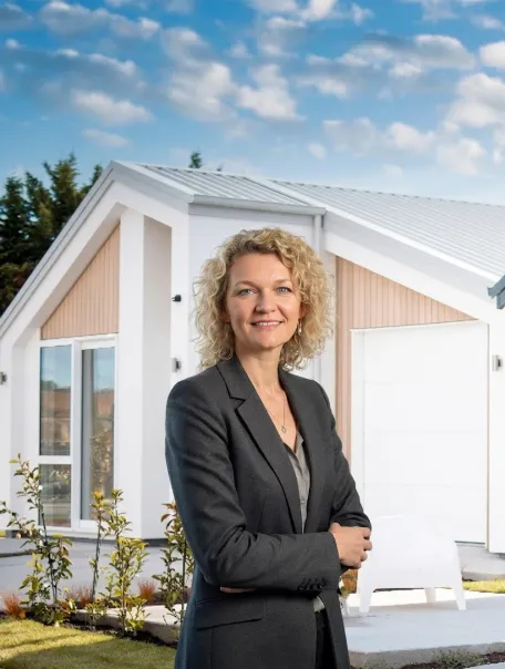 Smiling woman with curly blonde hair in a dark blazer standing with arms crossed in front of a modern white house with large windows and a sunlit garden.