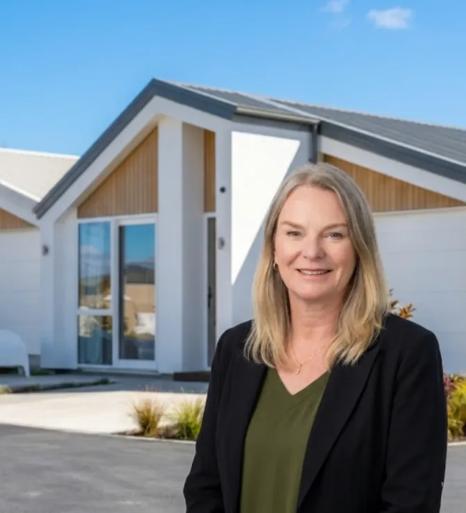 Smiling middle-aged woman with blonde hair wearing a black blazer and olive green top standing outside in front of modern white houses under a clear blue sky.