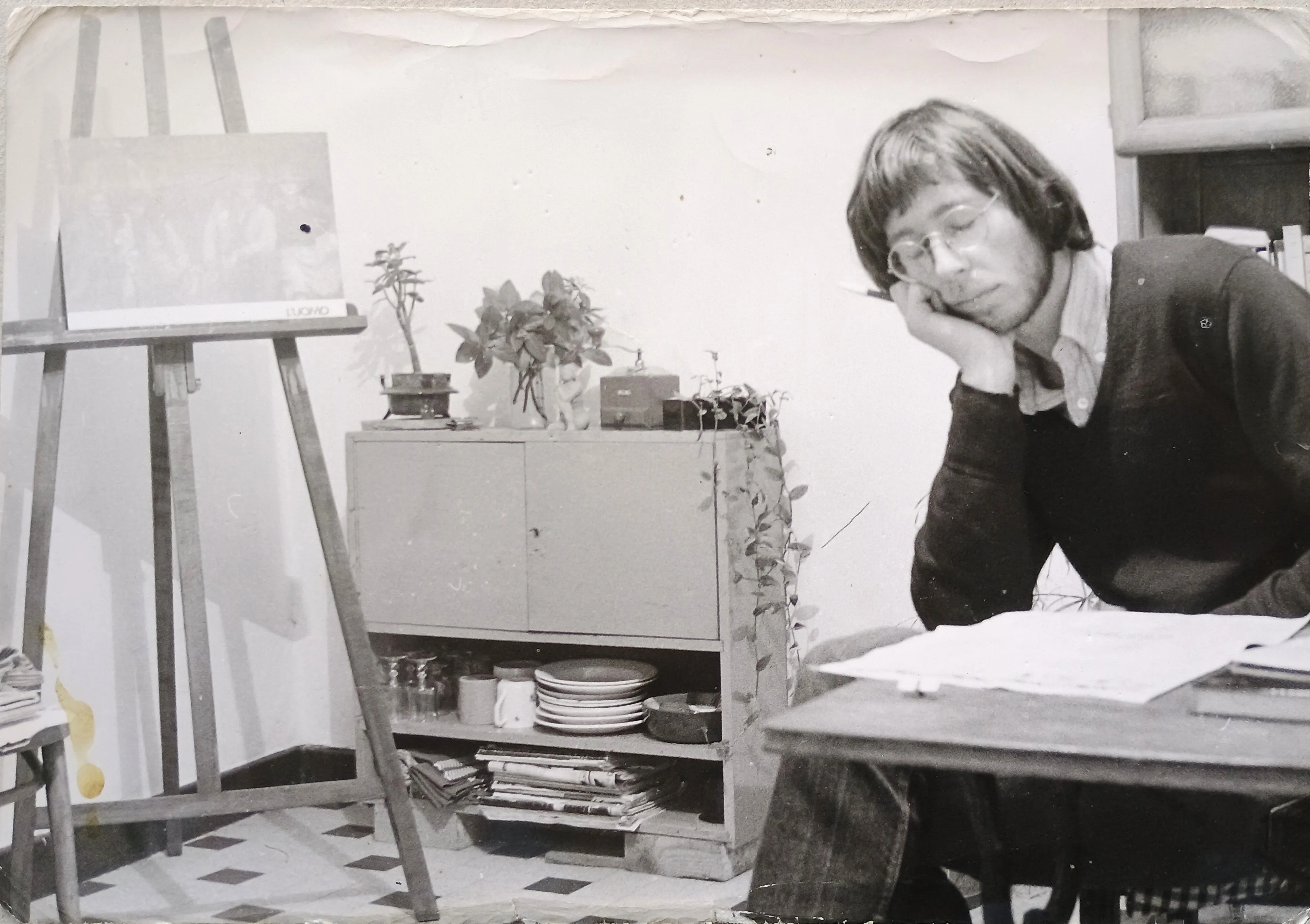 Black and white photo of a man with glasses resting his head on his hand while seated at a table with papers, next to an easel and a cabinet with plants and dishes.