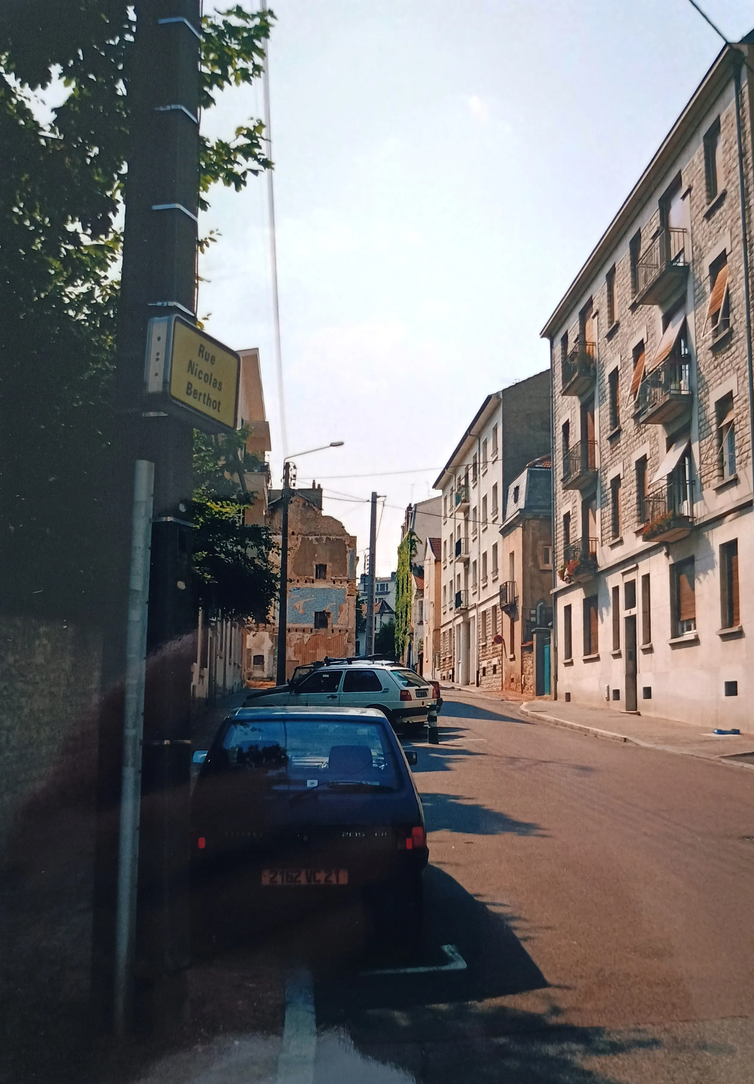 Sunny view of Rue Nicolas Berthot street with parked cars and old residential buildings with balconies and awnings.