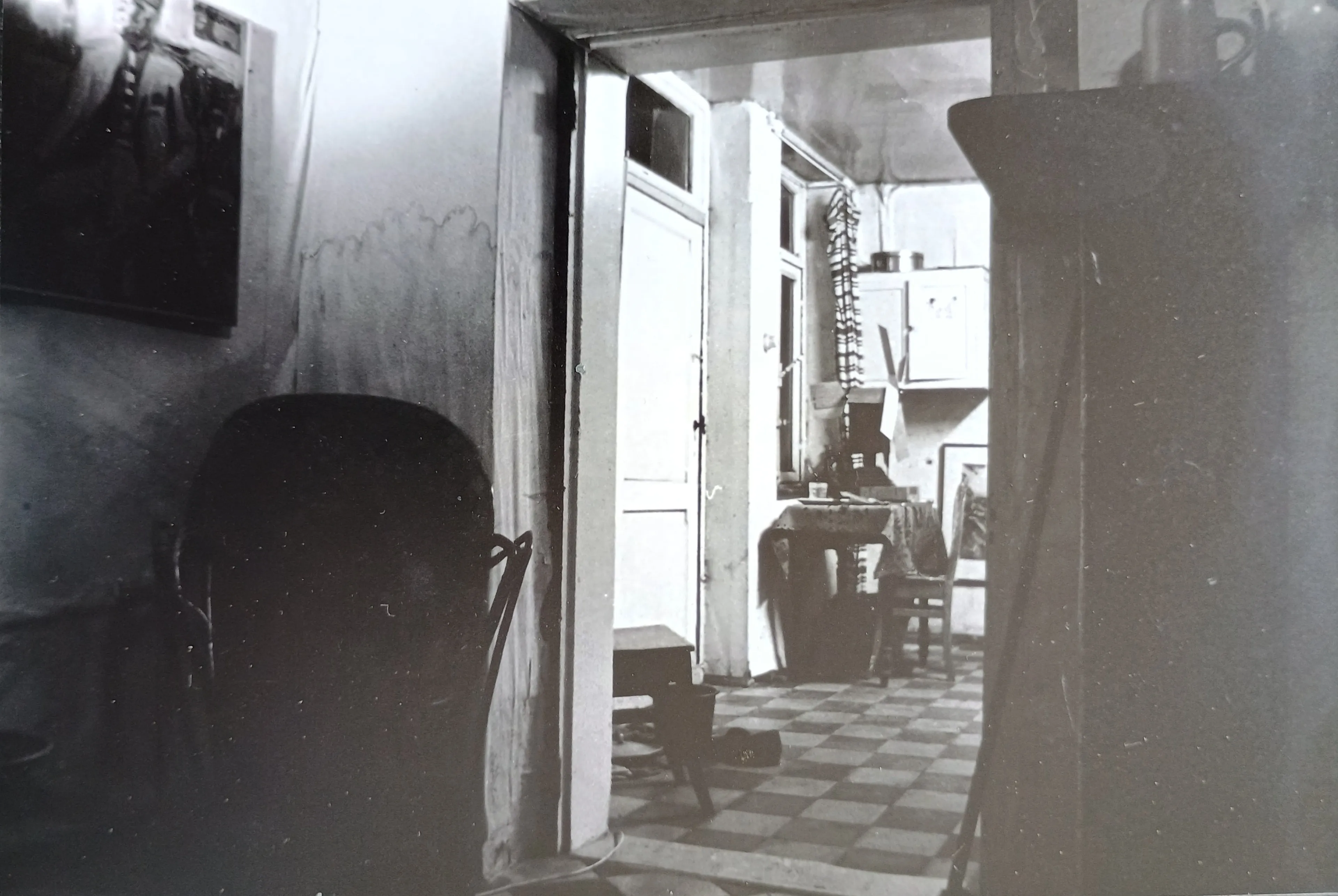 Black and white photo of a dimly lit room with checkered floor, wooden chairs, a table with a cloth, and a door in the background.