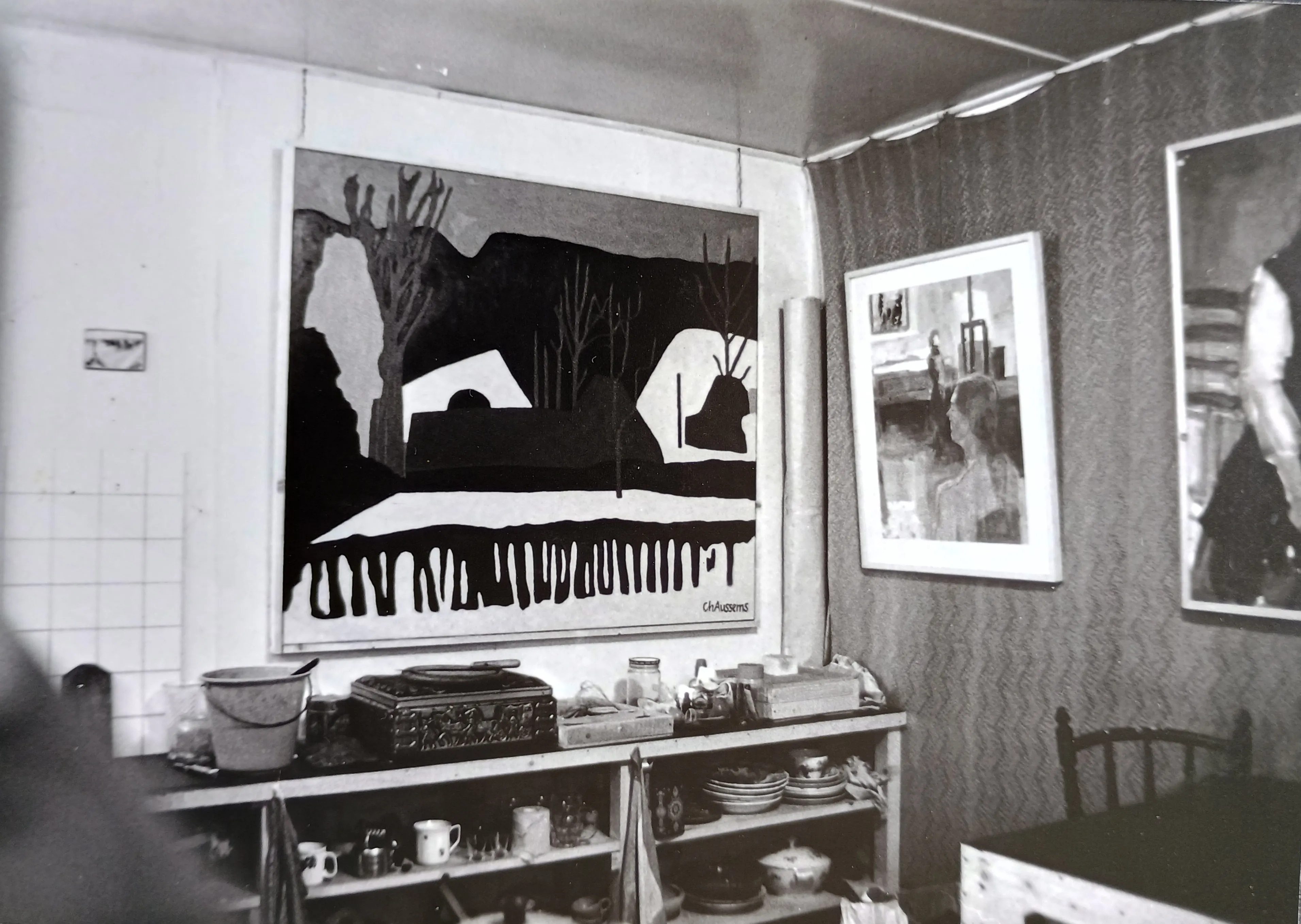 Black and white photo of a room with patterned wallpaper and three framed paintings on the walls, a wooden chair, and a shelf holding various kitchen items including cups and bowls.
