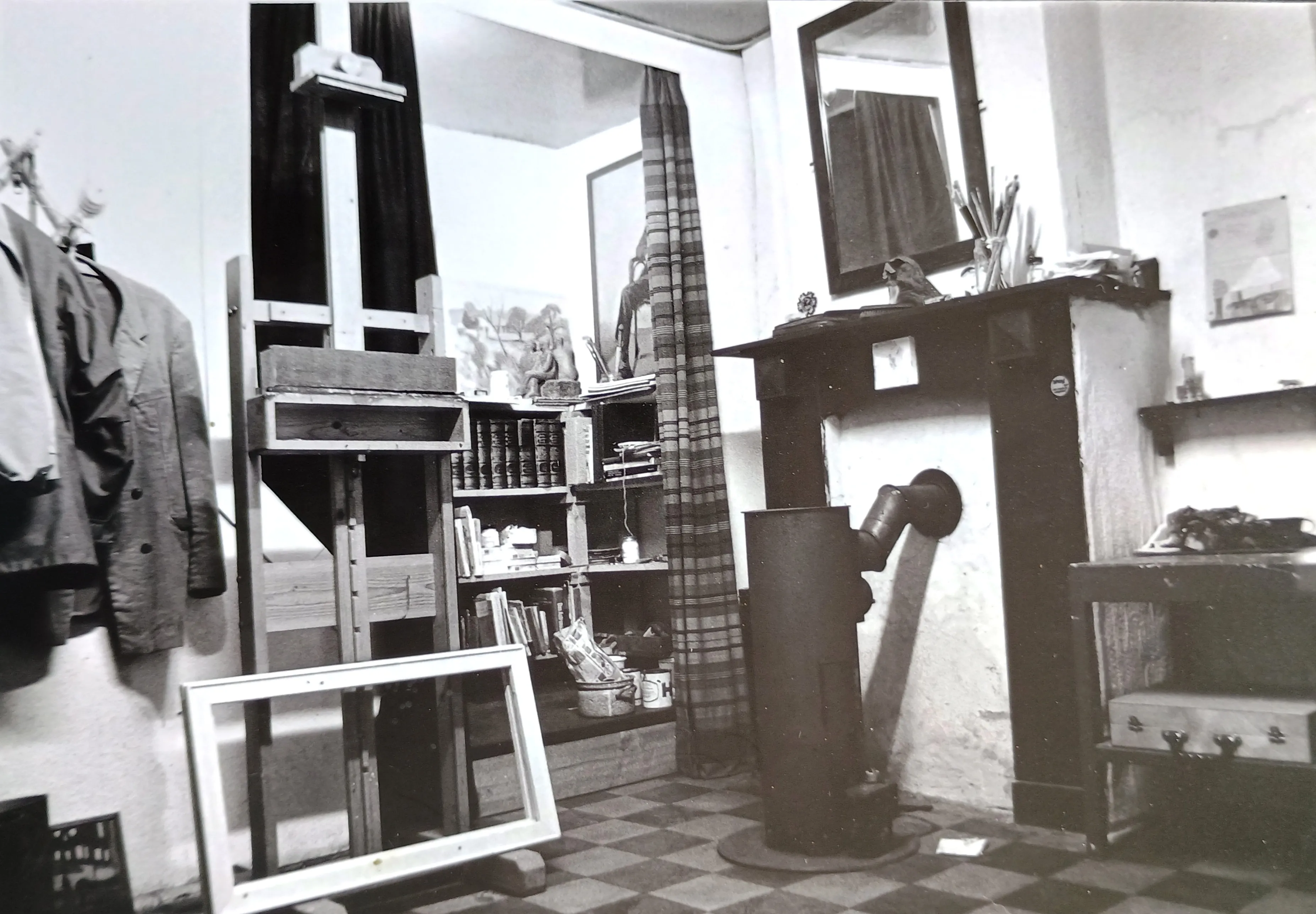 Black and white photo of an artist's studio with an easel, empty picture frame, bookshelf with books and art supplies, wood stove, and hanging coats.
