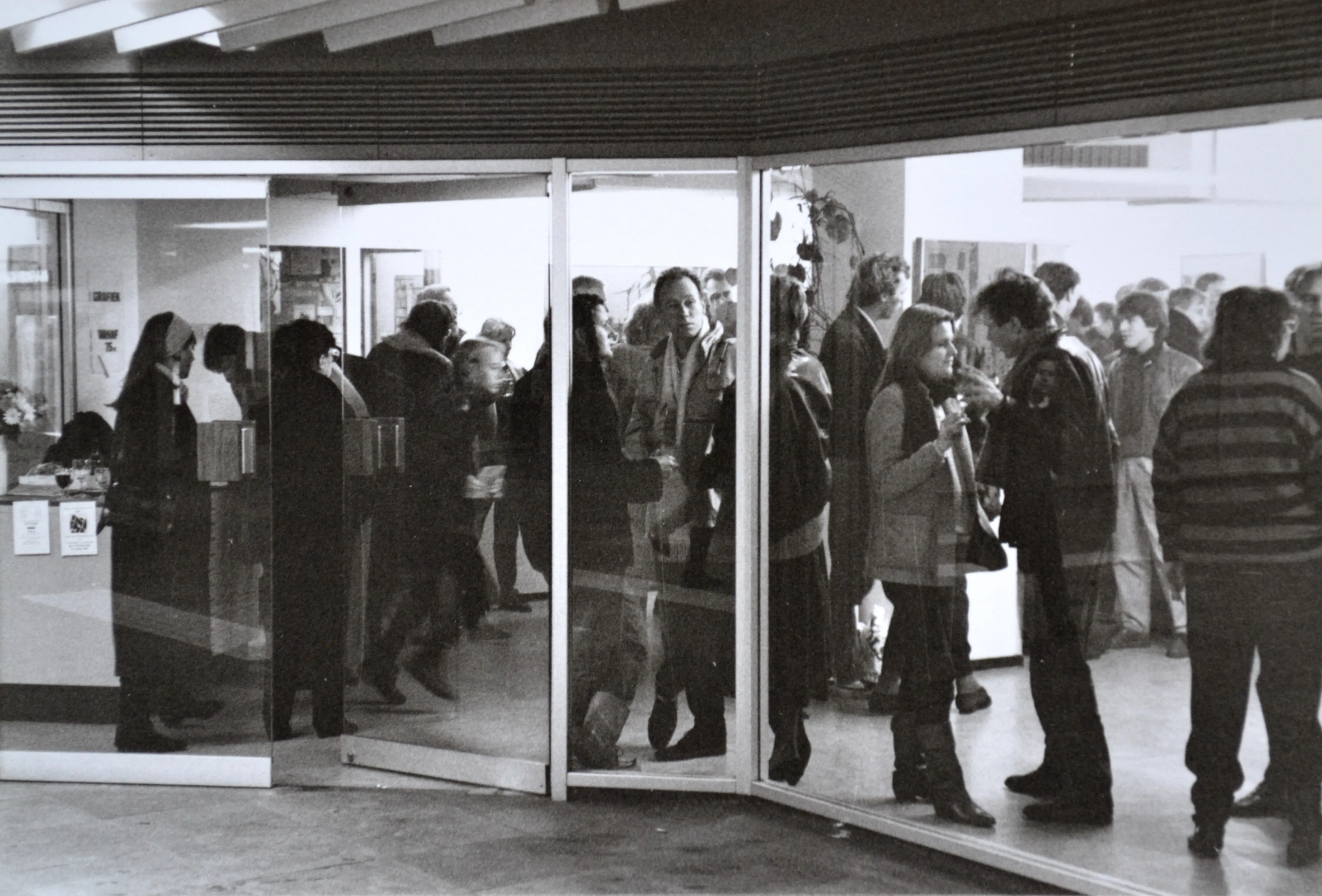 Black and white photo of a crowd of people gathered inside a building near glass doors, engaging in conversation.