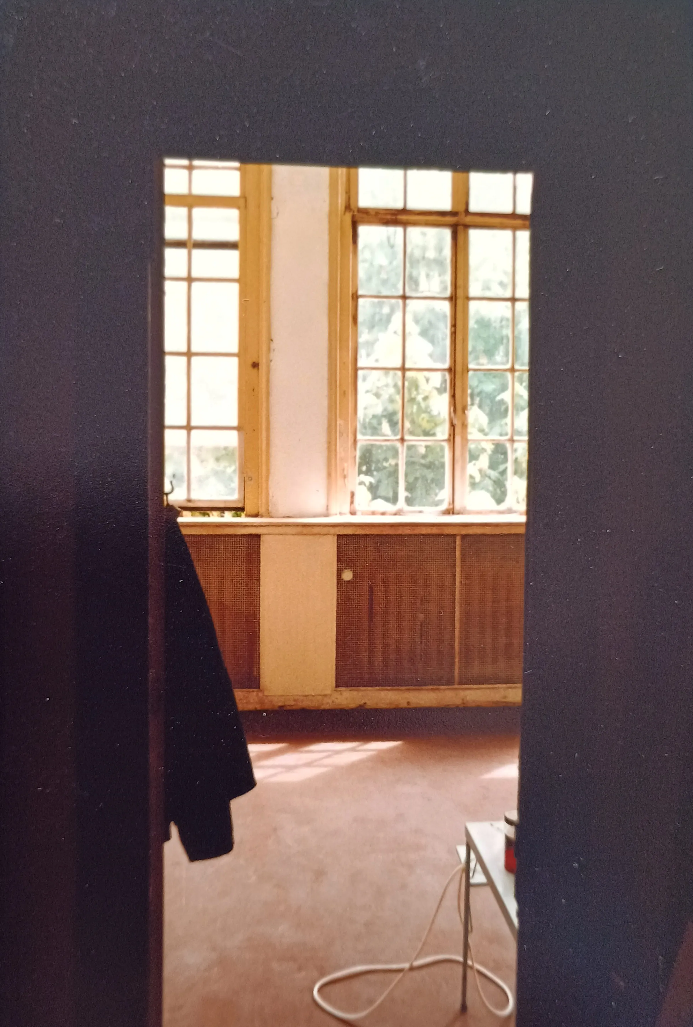 Sunlit room with large wooden-framed windows, a tiled radiator cover, and a white table with a red can and electric cord on the floor.