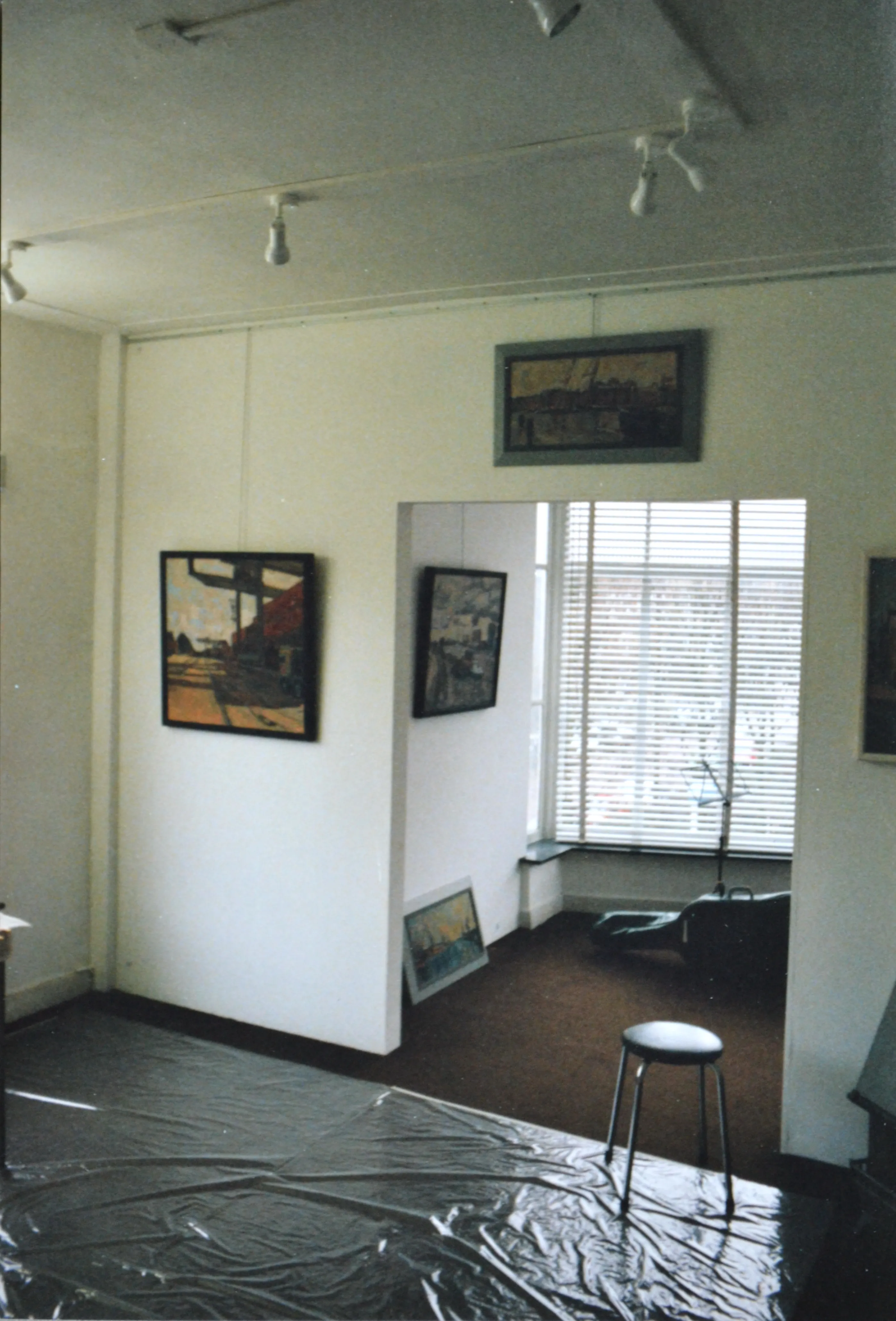 Interior of an art gallery room with paintings on white walls, a metal stool on a shiny floor, and a large window with blinds in the background.