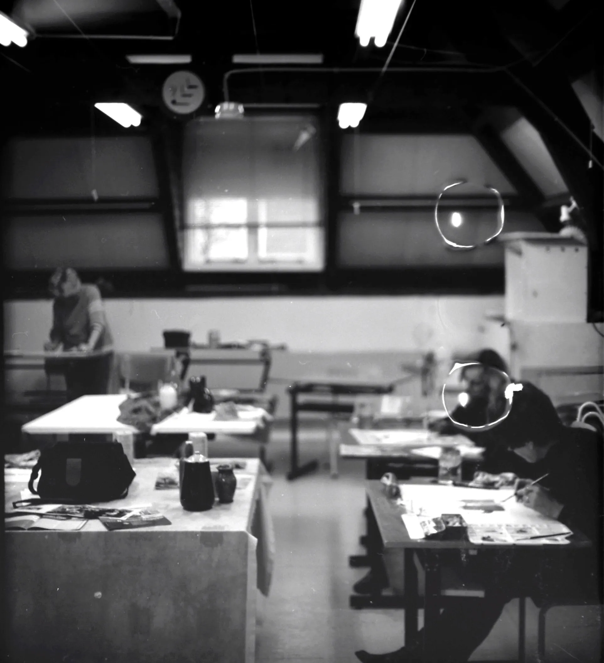 Black and white photo of three people working at tables in a dimly lit room with high ceilings and large windows.