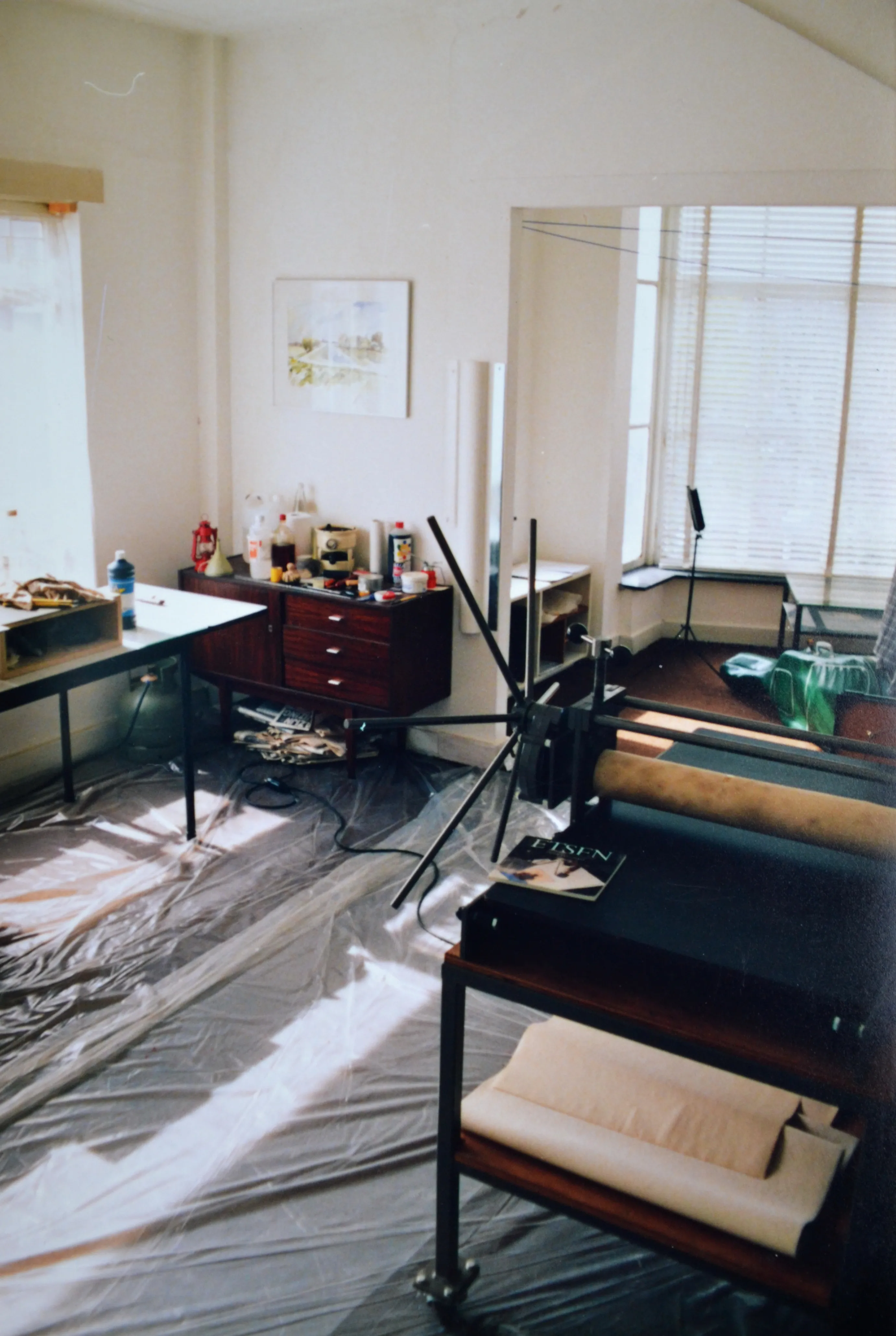 Art studio with sunlight on plastic-covered floor, scanograph equipment, wooden cabinet with art supplies, and large windows with blinds.