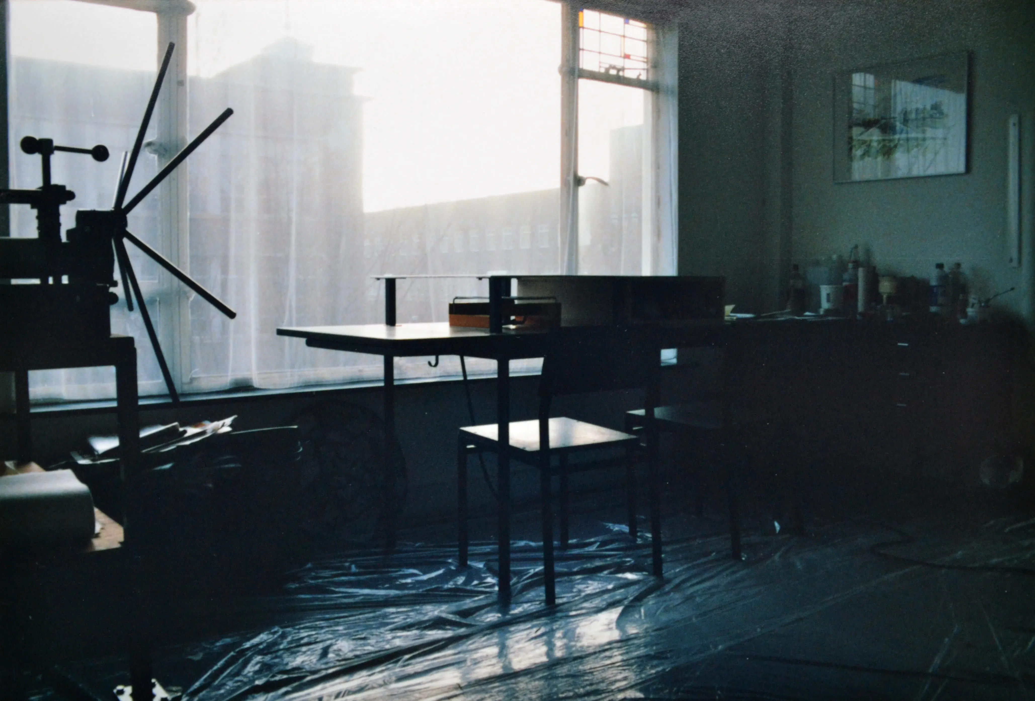Dimly lit workshop with a table, chairs, art supplies, and sheer curtains covering large windows.