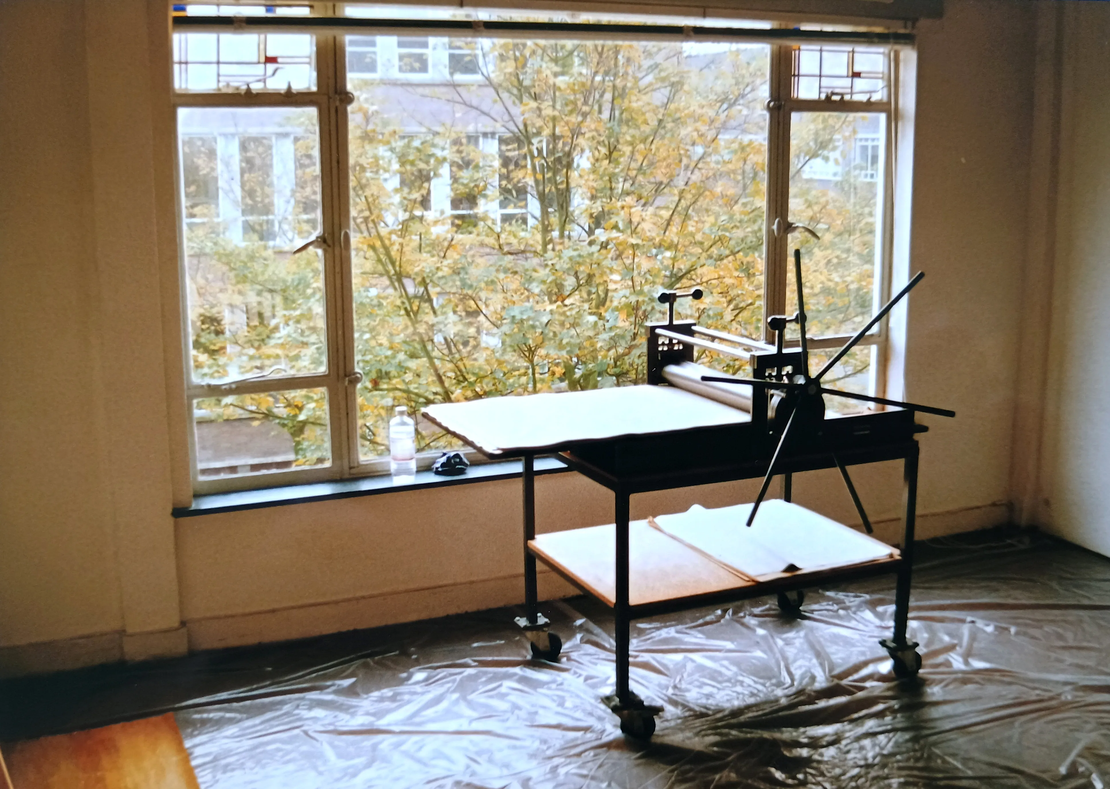 Etching press machine on wheels positioned near a large window with autumn trees outside.