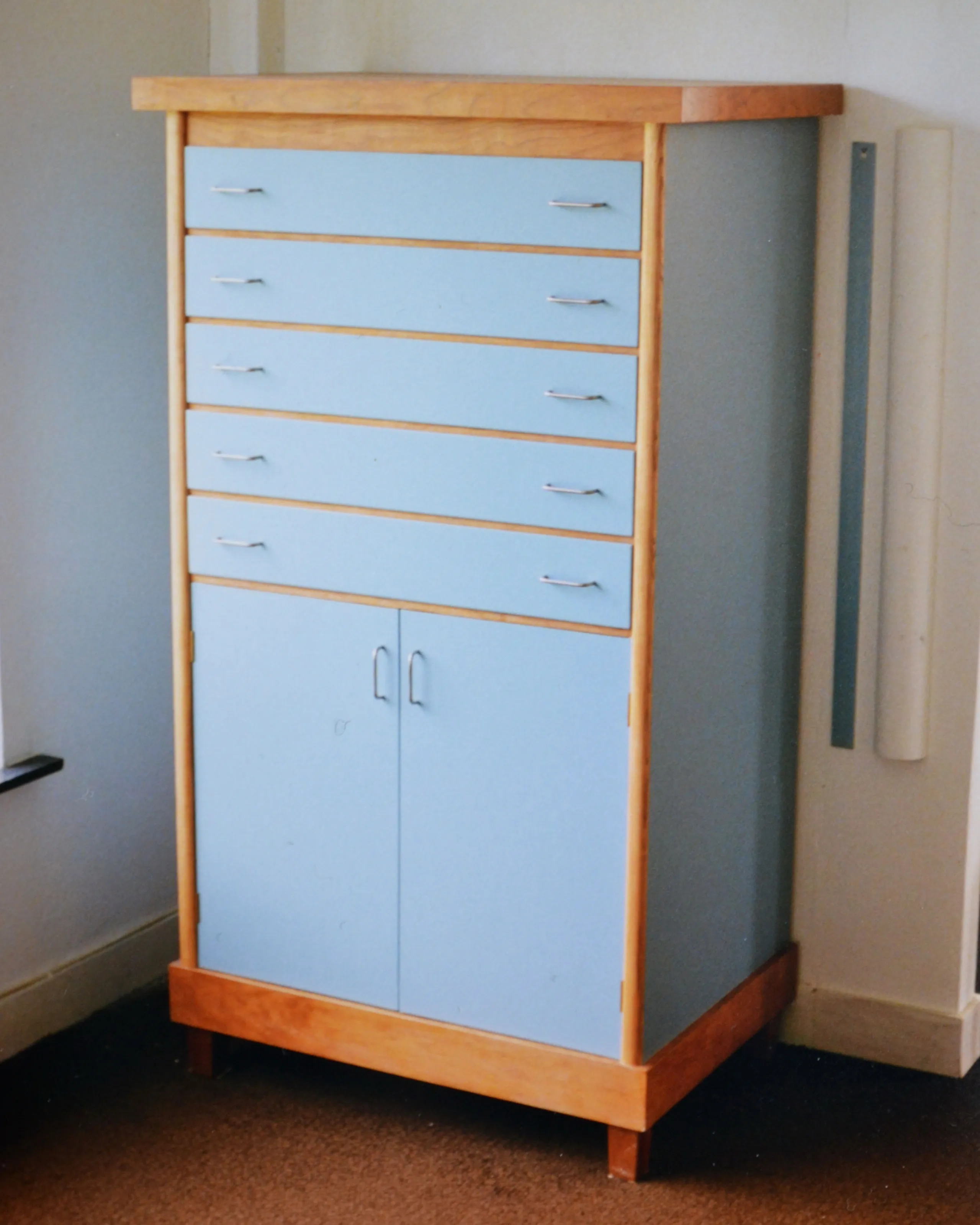 Light blue cabinet with five drawers and two doors, framed with light wood, standing on a brown carpeted floor.