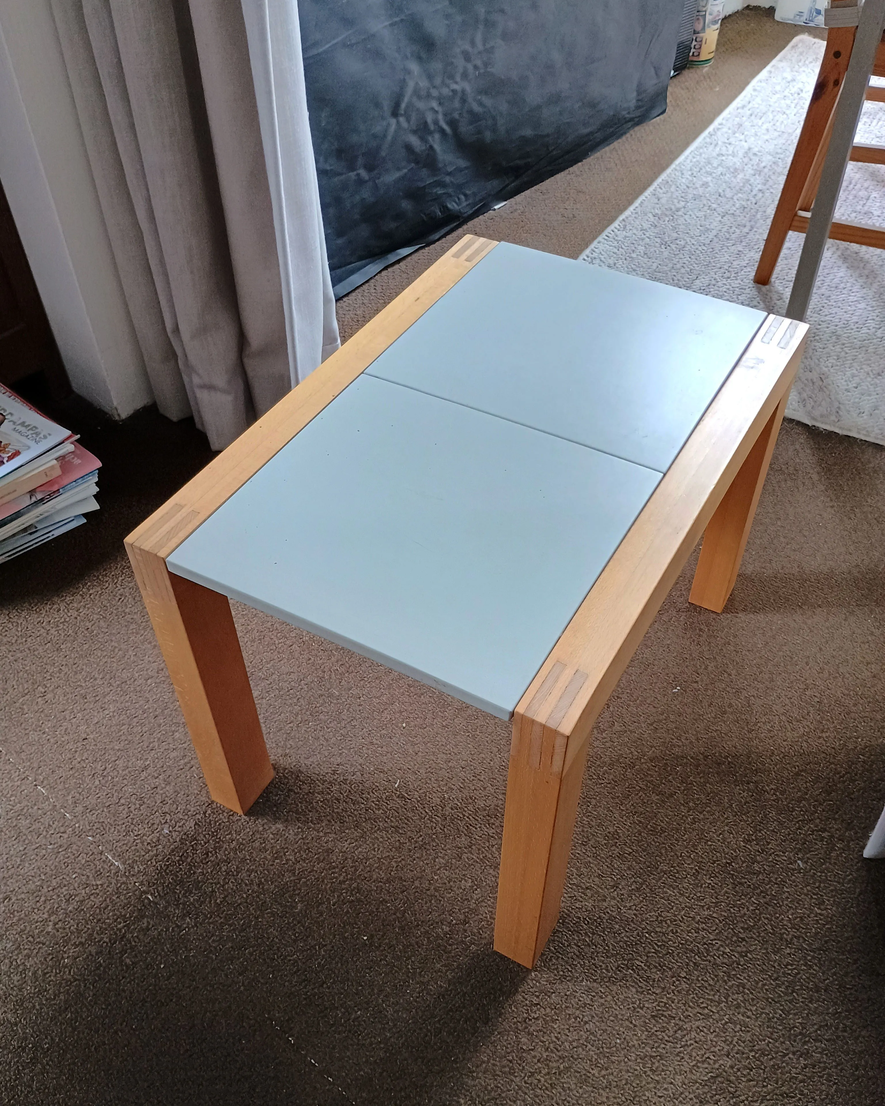 Small rectangular wooden side table with light gray tiled top, placed on brown carpet indoors.