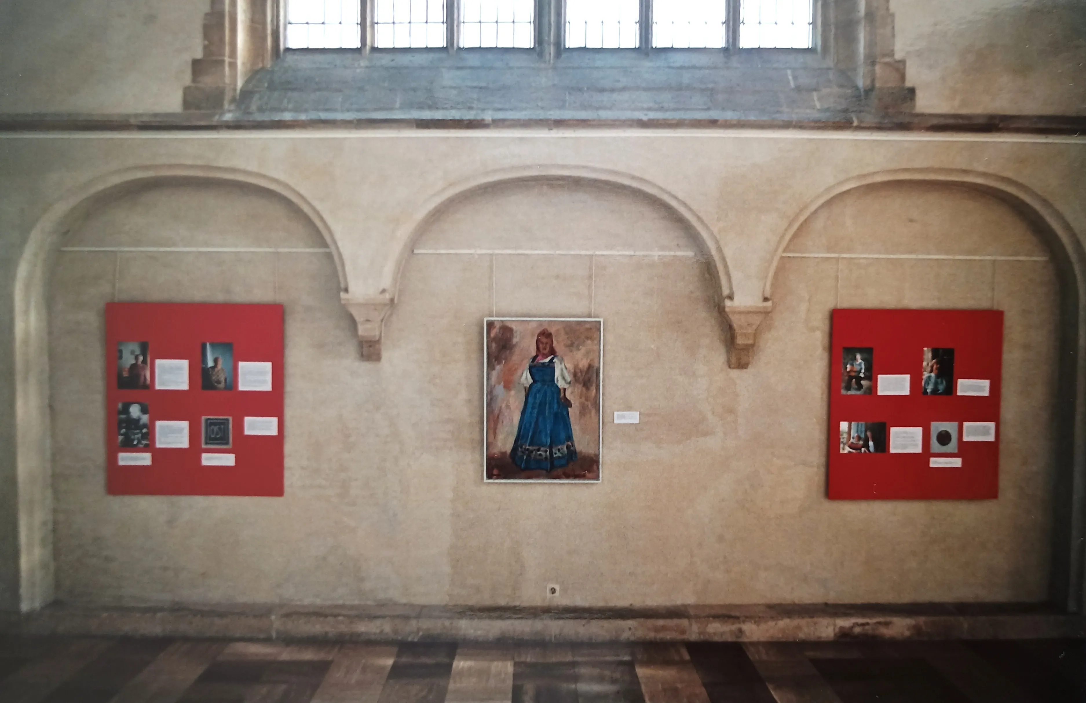 Exhibition wall with three arched niches featuring a central portrait of a woman in traditional blue dress and two red panels with photos and text on either side.