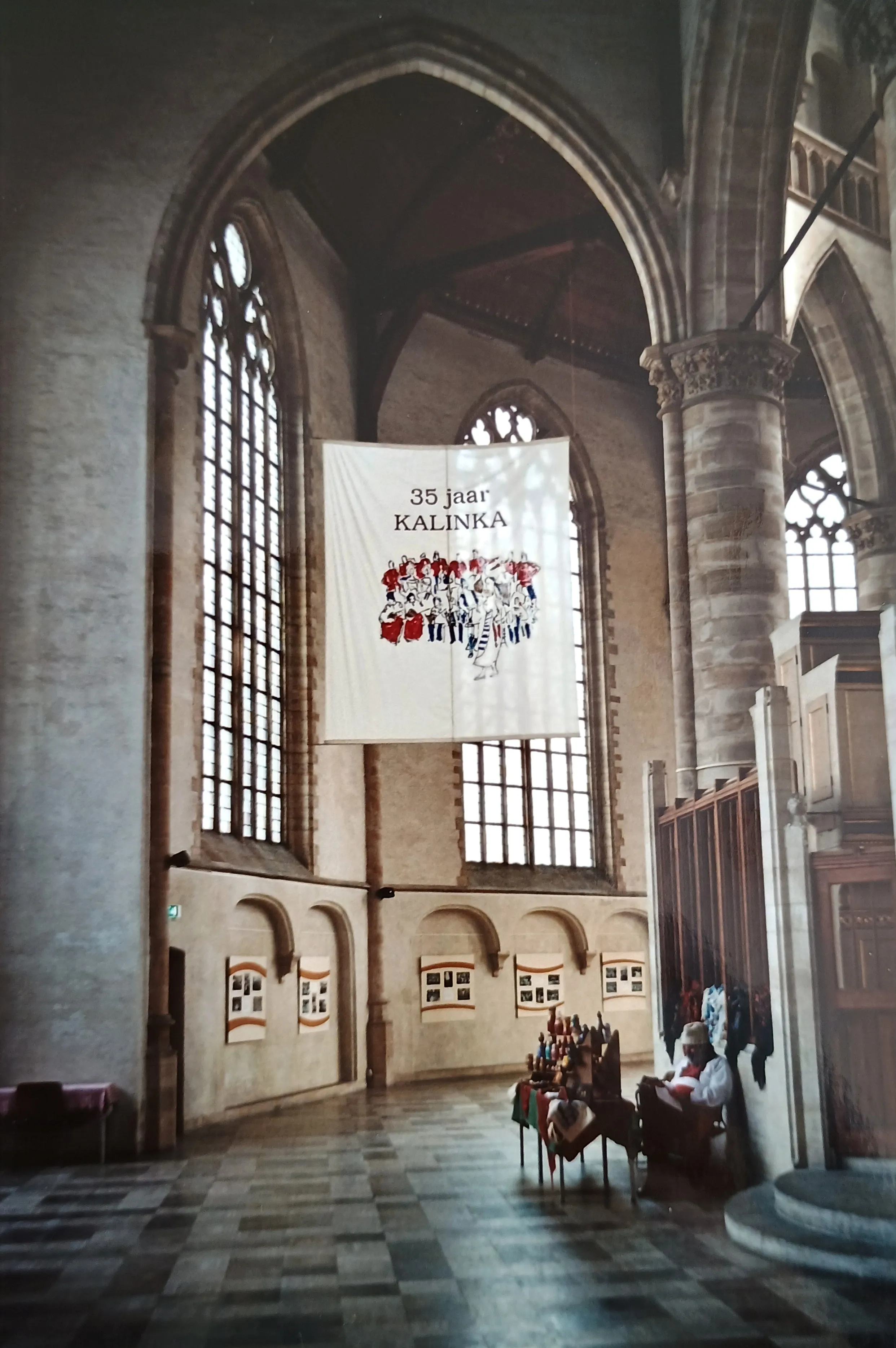 Interior of a historic church with tall arched windows, a large hanging banner celebrating 35 years of Kalinka, and a person seated near a table displaying colorful items.