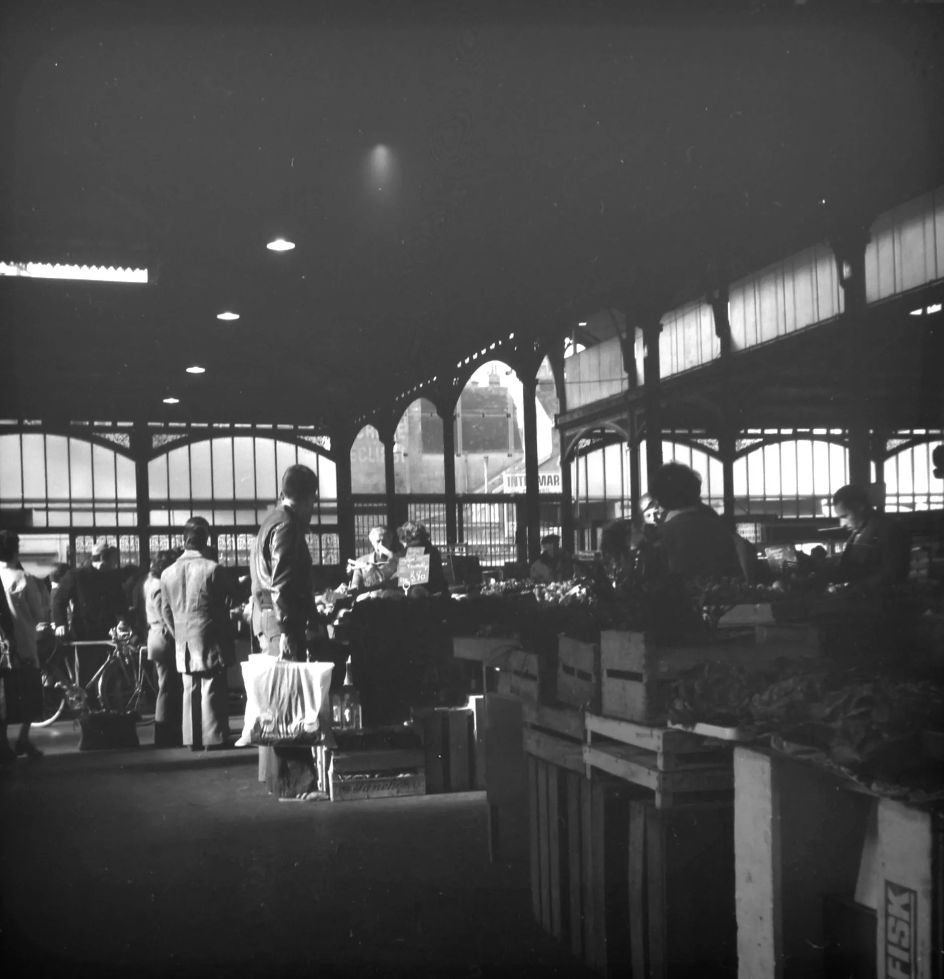 Black and white photo of people shopping at an indoor market with produce crates and high arched windows.